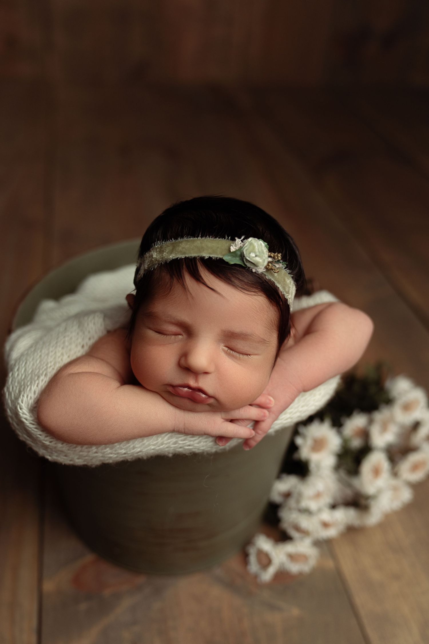 Sleeping newborn baby girl in green bucket with floral headband and white flowers on wood floor.