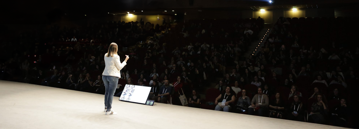 Speaker presenting on stage to a large audience in a packed auditorium during a conference event.