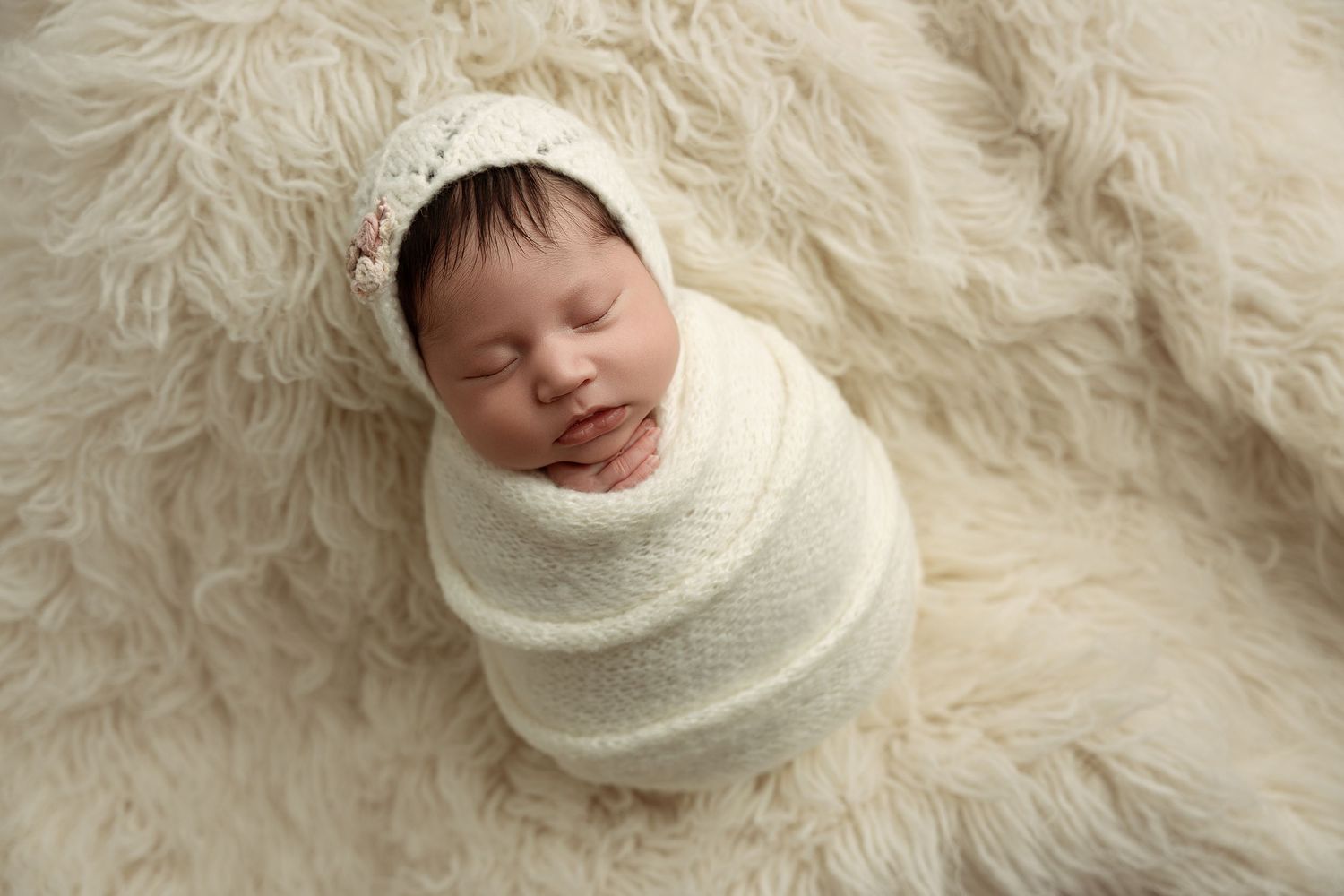 Newborn baby swaddled in cream wrap with knit bonnet, sleeping peacefully on fluffy white fur blanket.