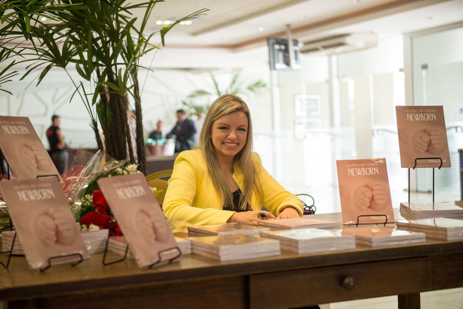 Smiling blonde woman in yellow jacket signing books at a wooden table display at an indoor book signing event.