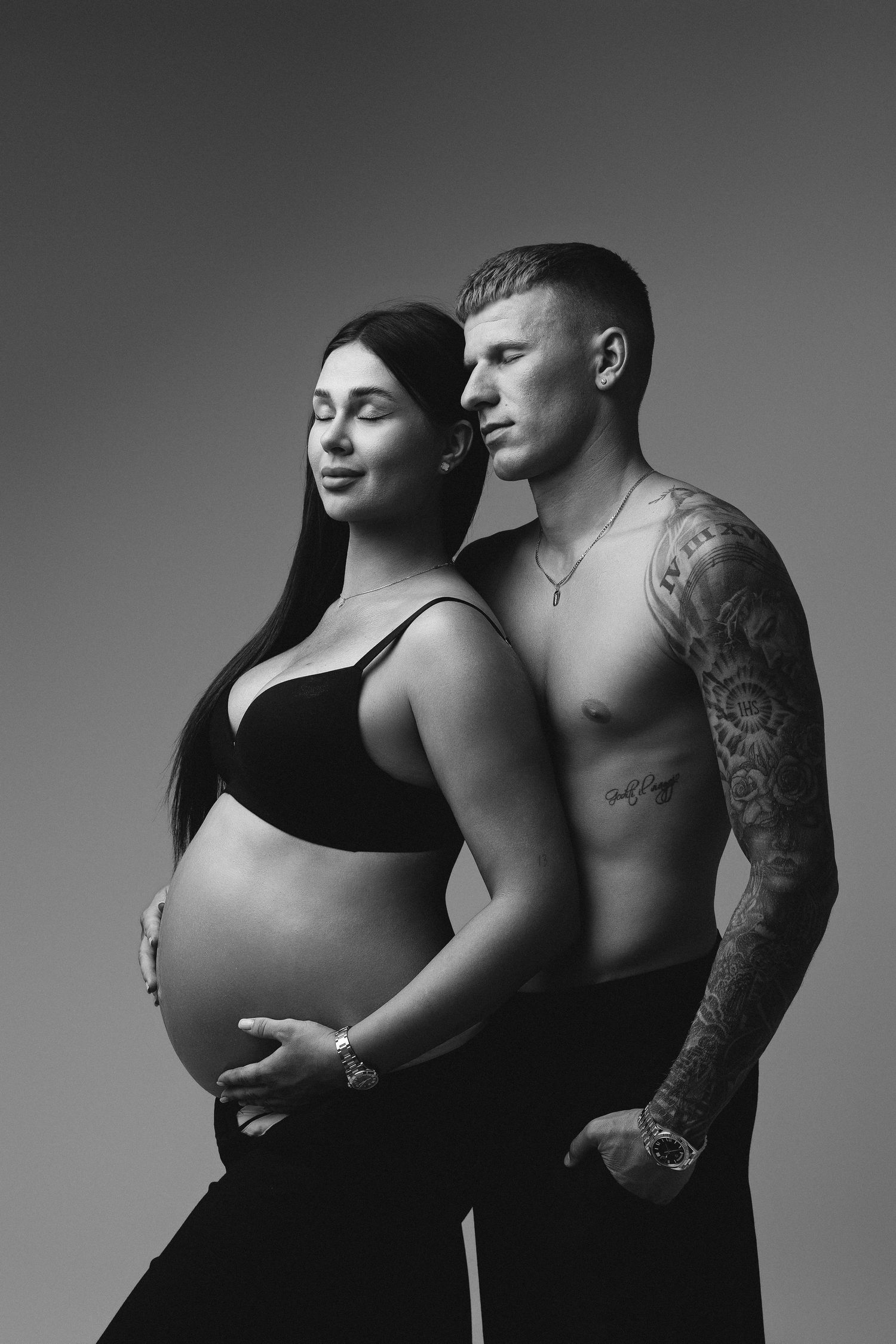Black and white maternity photo of pregnant woman in crop top with tattooed shirtless man posing together in studio.