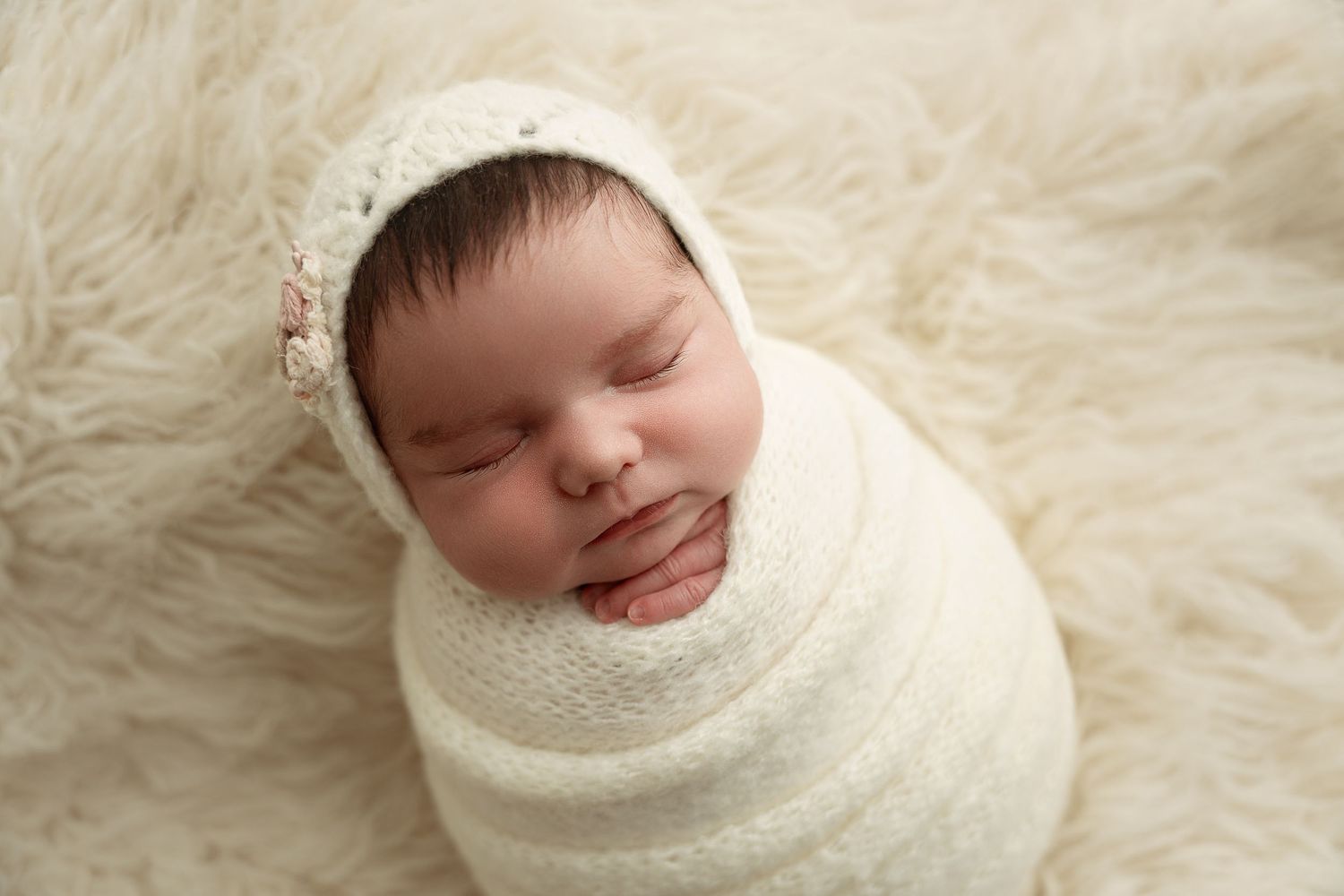 A sleeping newborn baby wrapped in a white blanket lies on soft white fur bedding. – newborn photographer Paloma Schell Houston TX