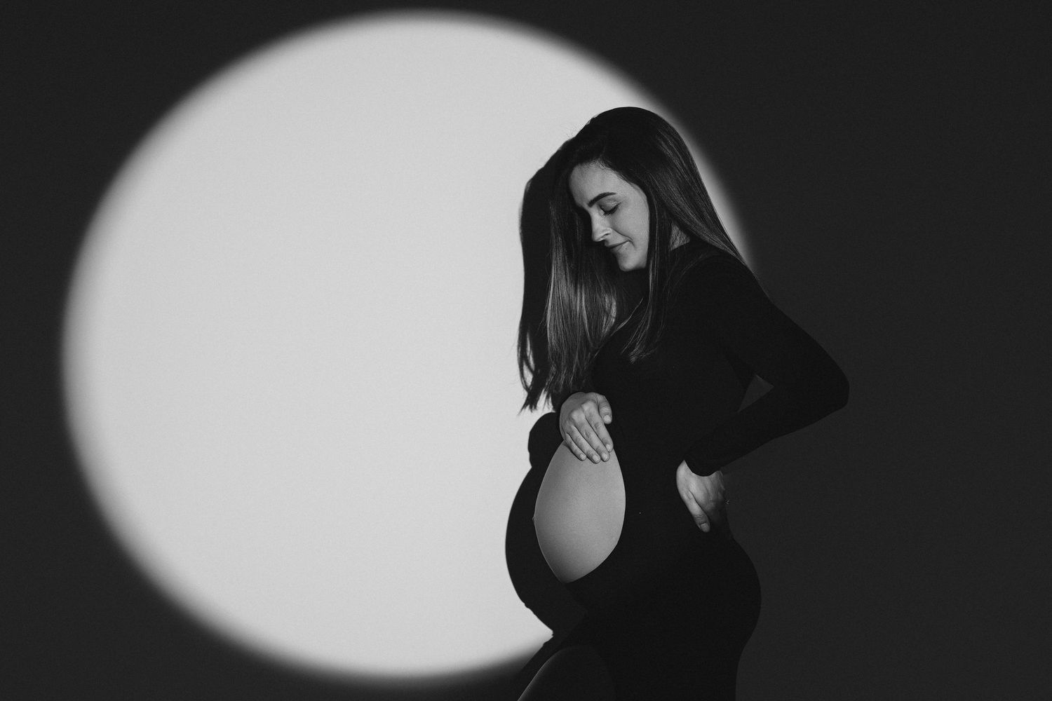 Black and white maternity photo of pregnant woman in black dress posed against circular light backdrop.