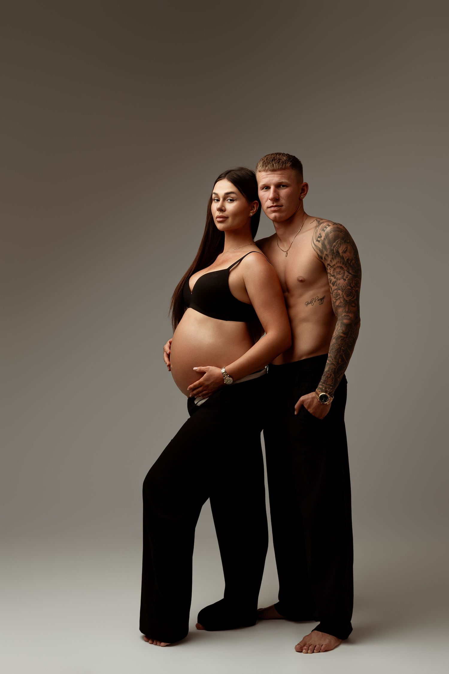 Pregnant woman in black bra and pants poses with tattooed shirtless man against gray background.