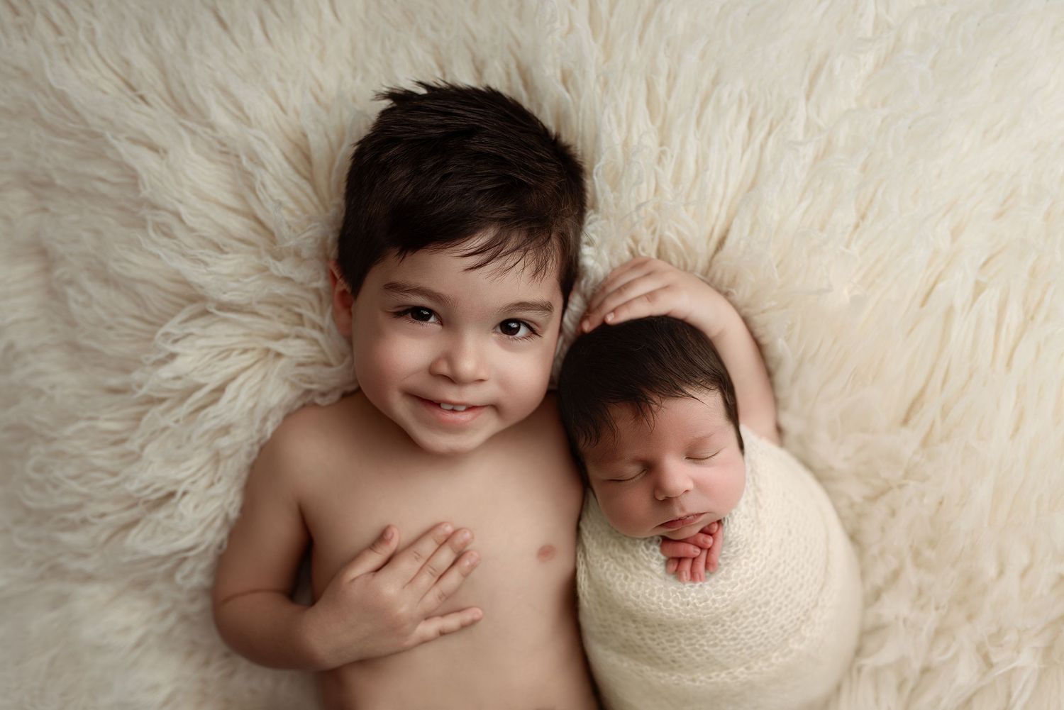 Smiling toddler boy lying beside sleeping newborn baby wrapped in cream blanket on fluffy white fur backdrop.