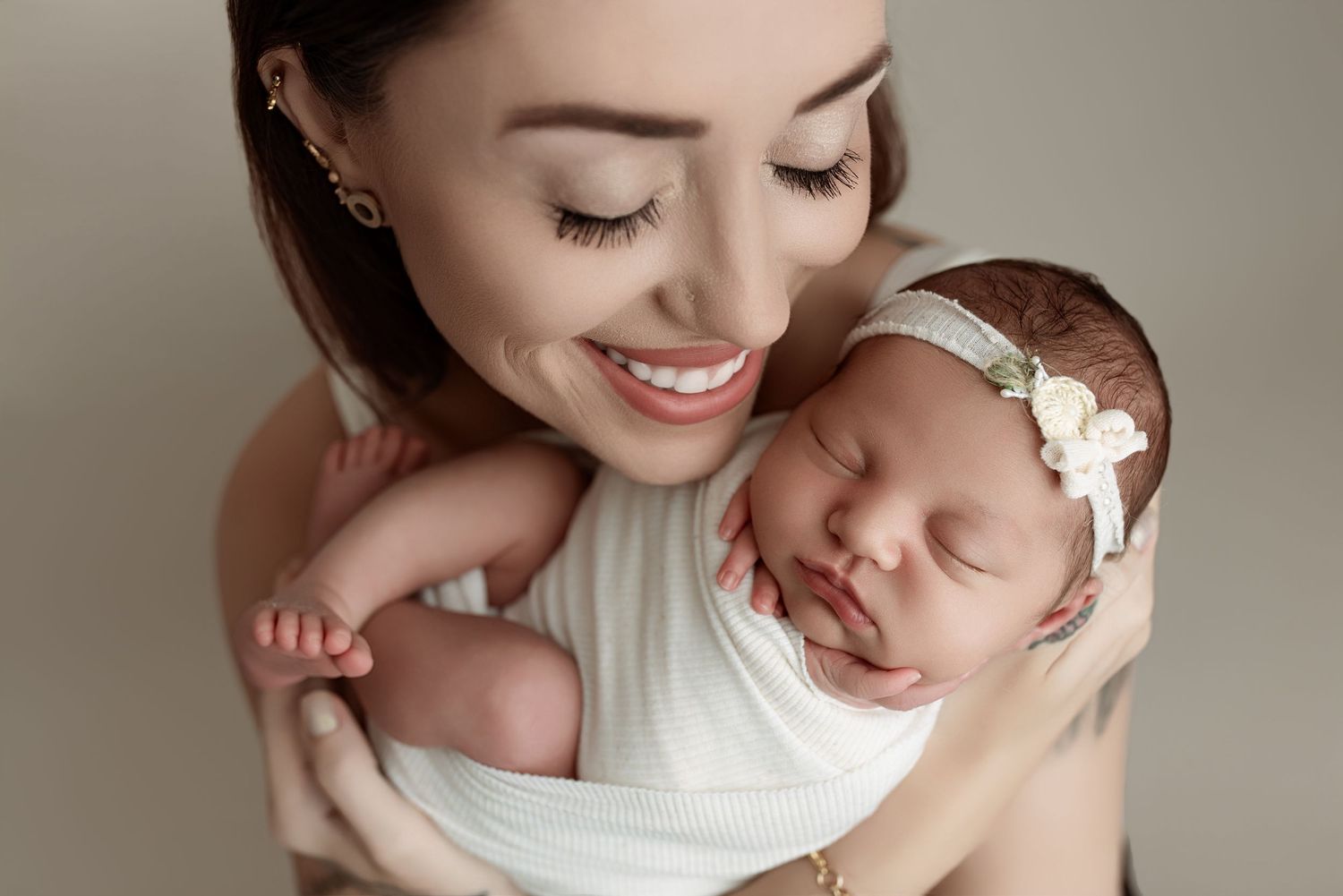 Smiling mother holds sleeping newborn baby in white wrap and floral headband against neutral background.