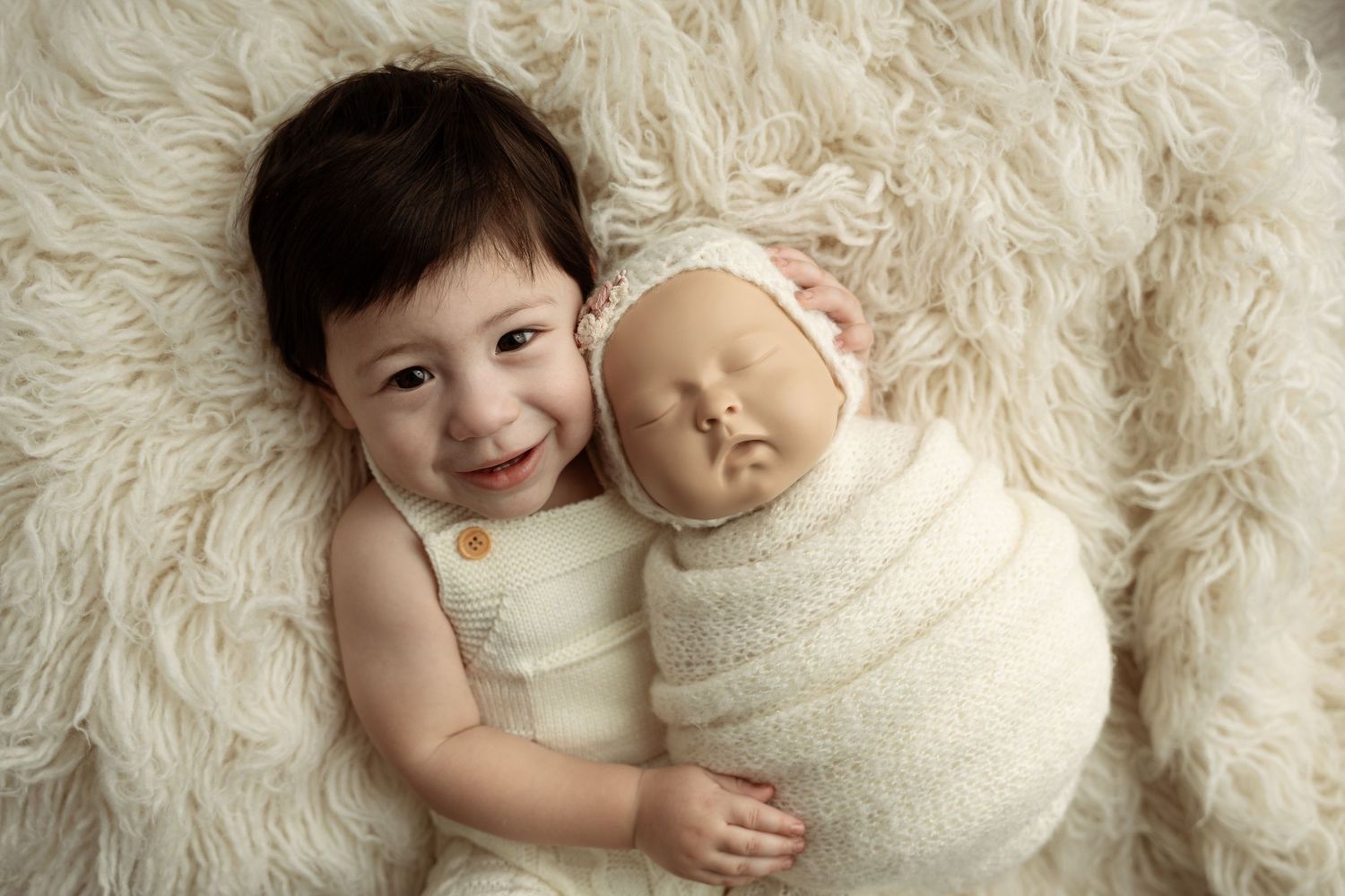 Two young children laying together on a soft white fur rug in matching cream-colored outfits– newborn photographer Paloma Schell Houston TX