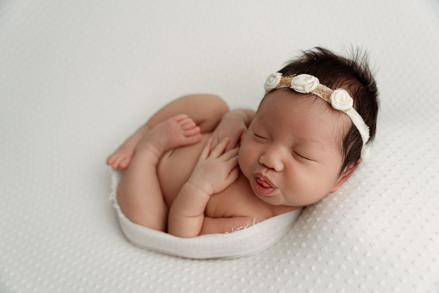 Sleeping newborn baby girl in white bowl wearing floral headband on white textured background.