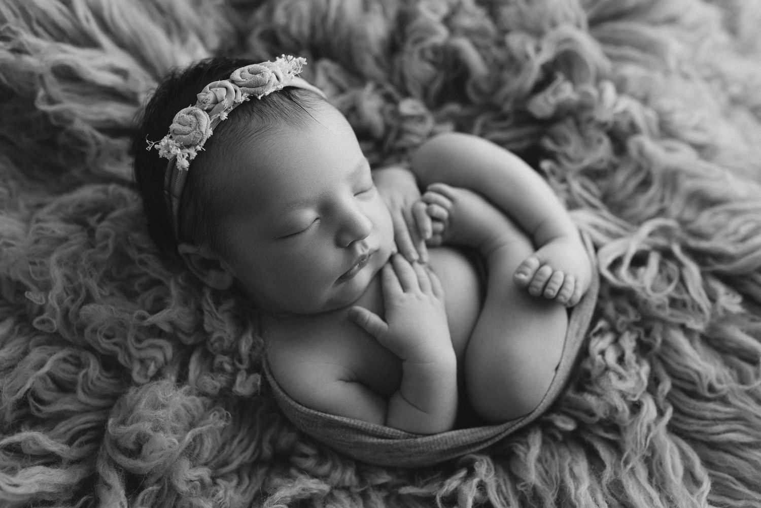 Black and white newborn photo of sleeping baby girl in bowl with floral headband on fluffy textured blanket.