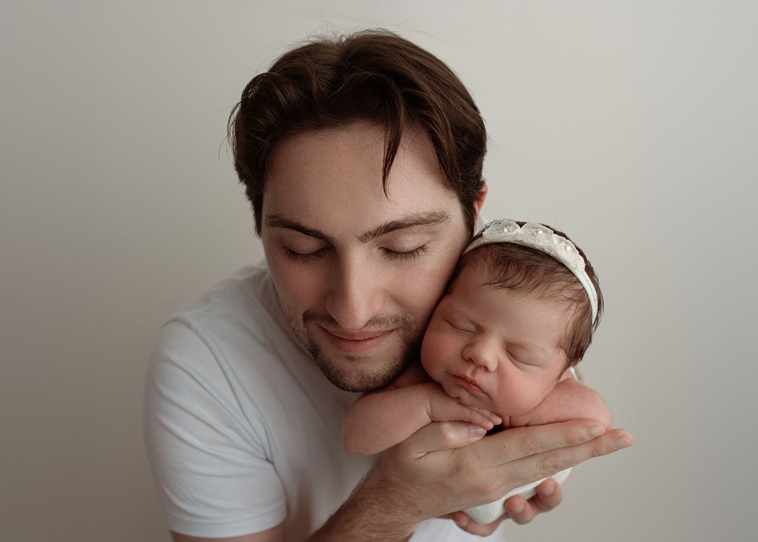 A smiling young man in a white t-shirt gently holds a sleeping newborn baby wearing a white headband in his hands.