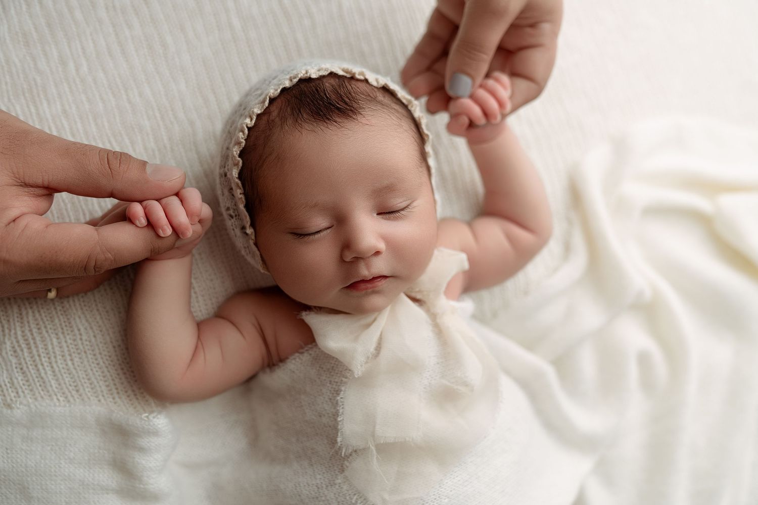 Sleeping newborn baby in white bonnet and outfit lying on soft white blanket held by parent hands.