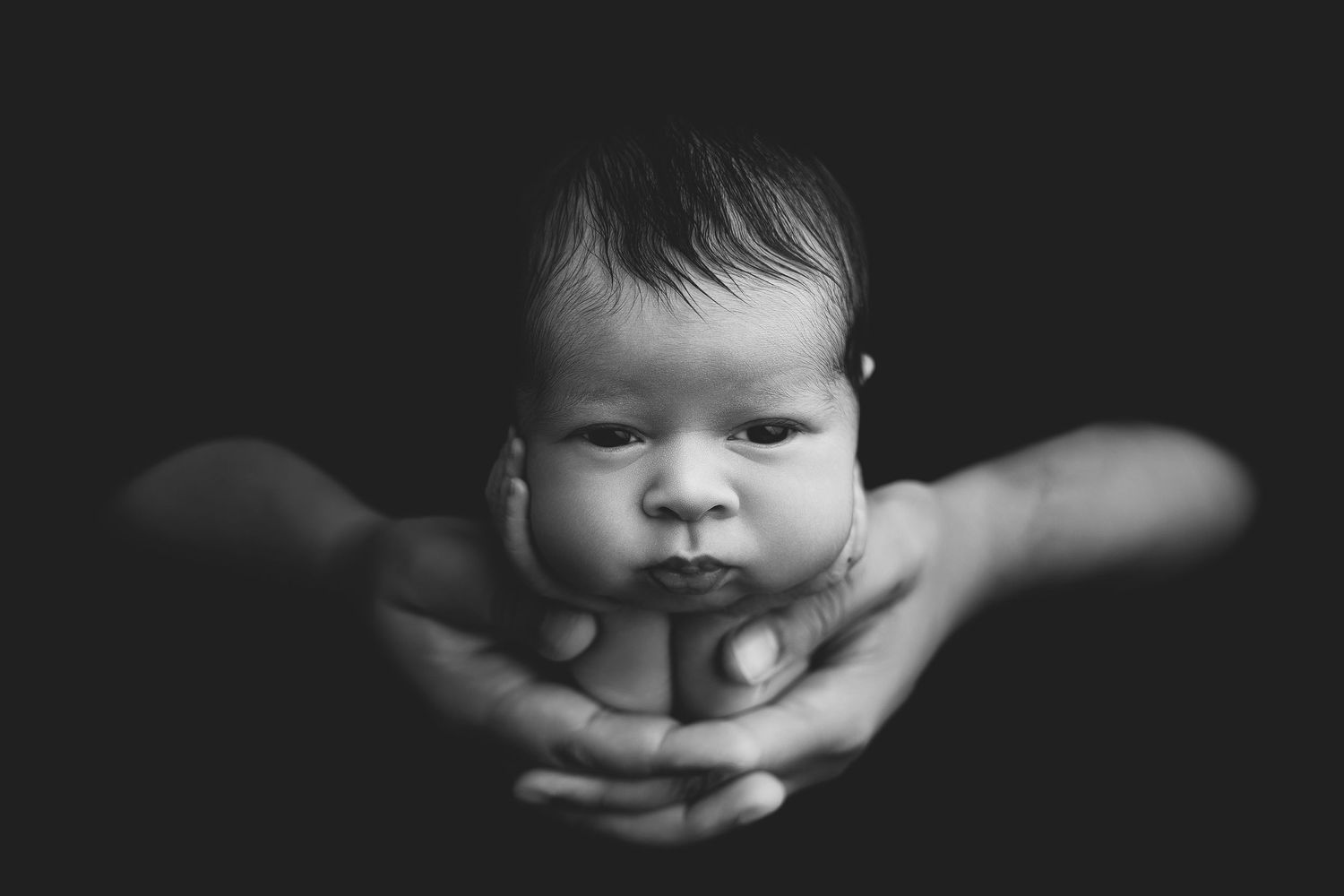 Black and white photo of a newborn baby cradled gently in adult hands against a dark background.