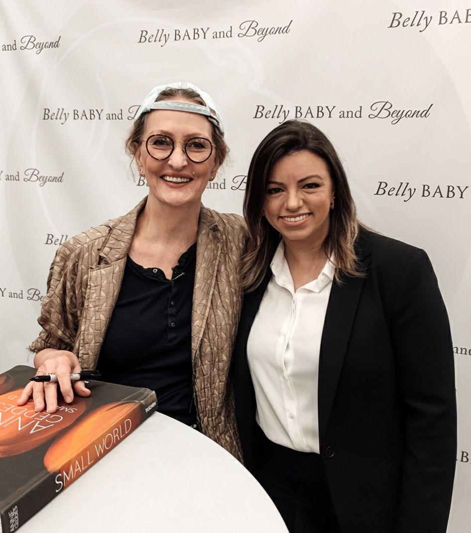 Two women posing together at a Belly Baby and Beyond event, one holding a book titled Small World.