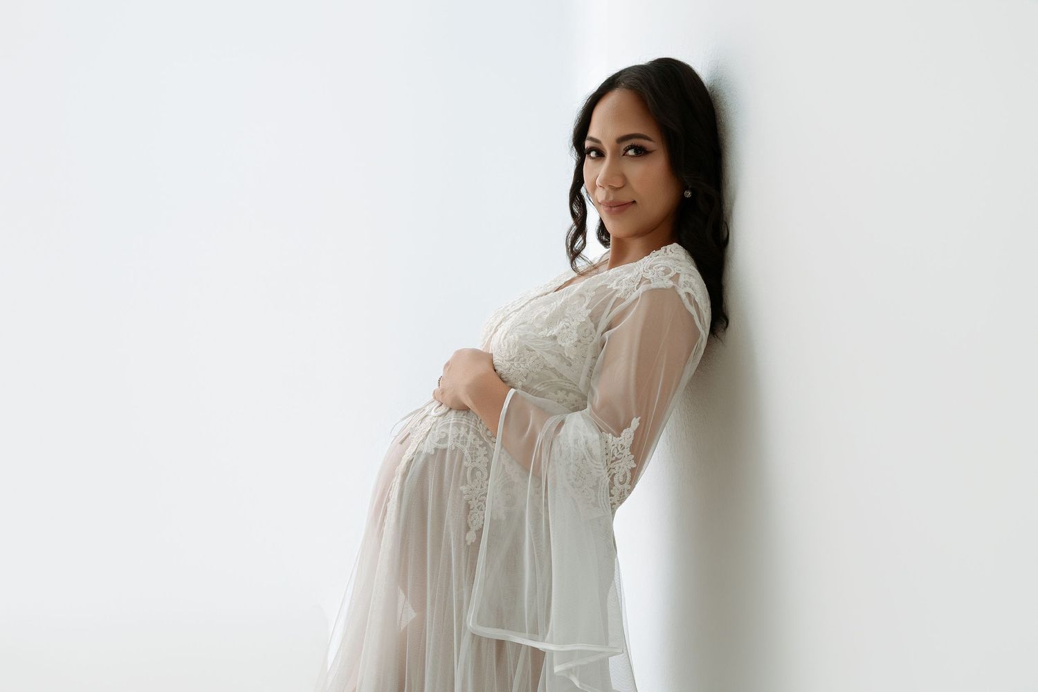 Woman in flowy white dress poses gracefully against white wall in minimalist fashion photoshoot.  maternity photographer Paloma Schell