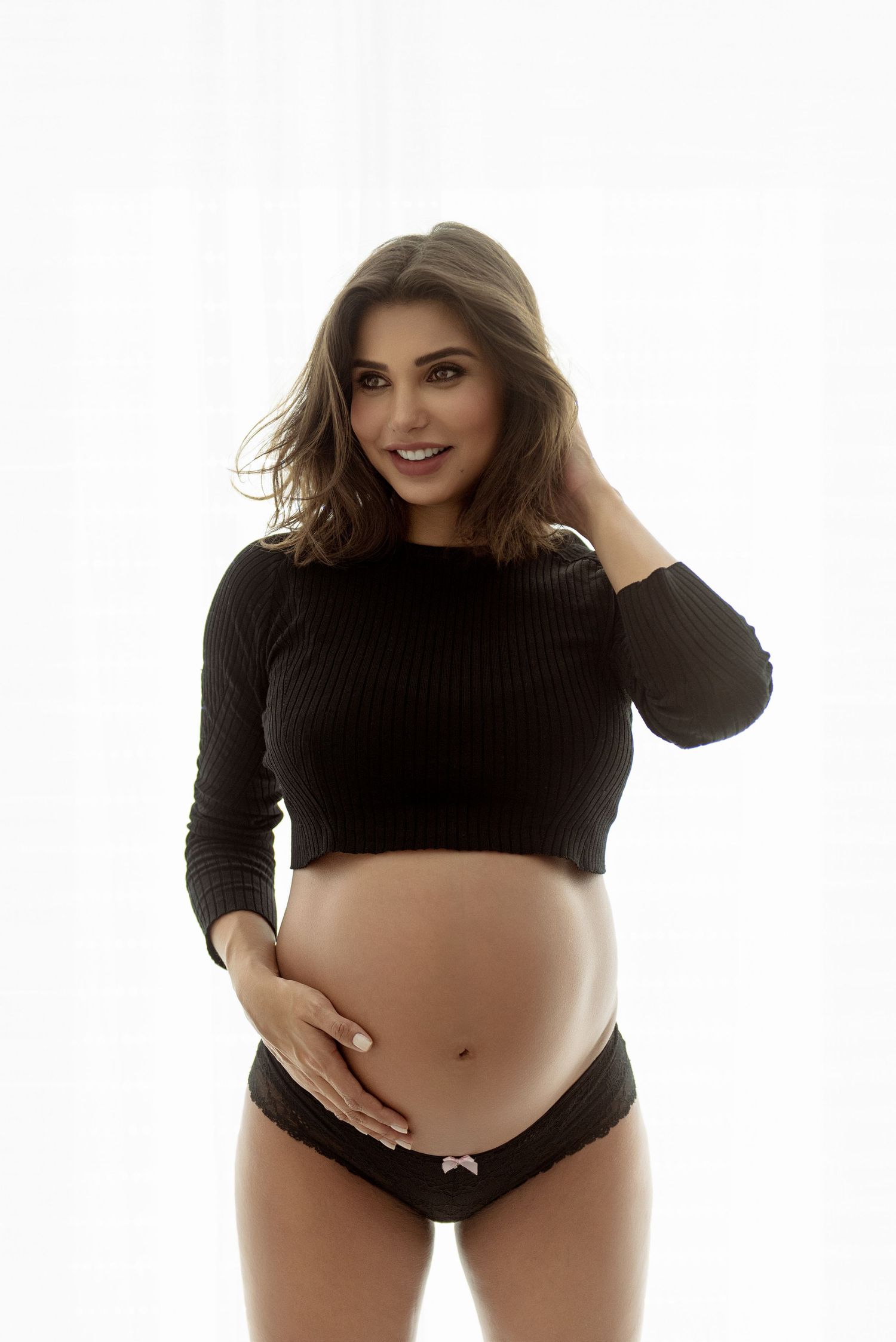Smiling pregnant woman in black crop top and lace underwear cradling her baby bump against white background.