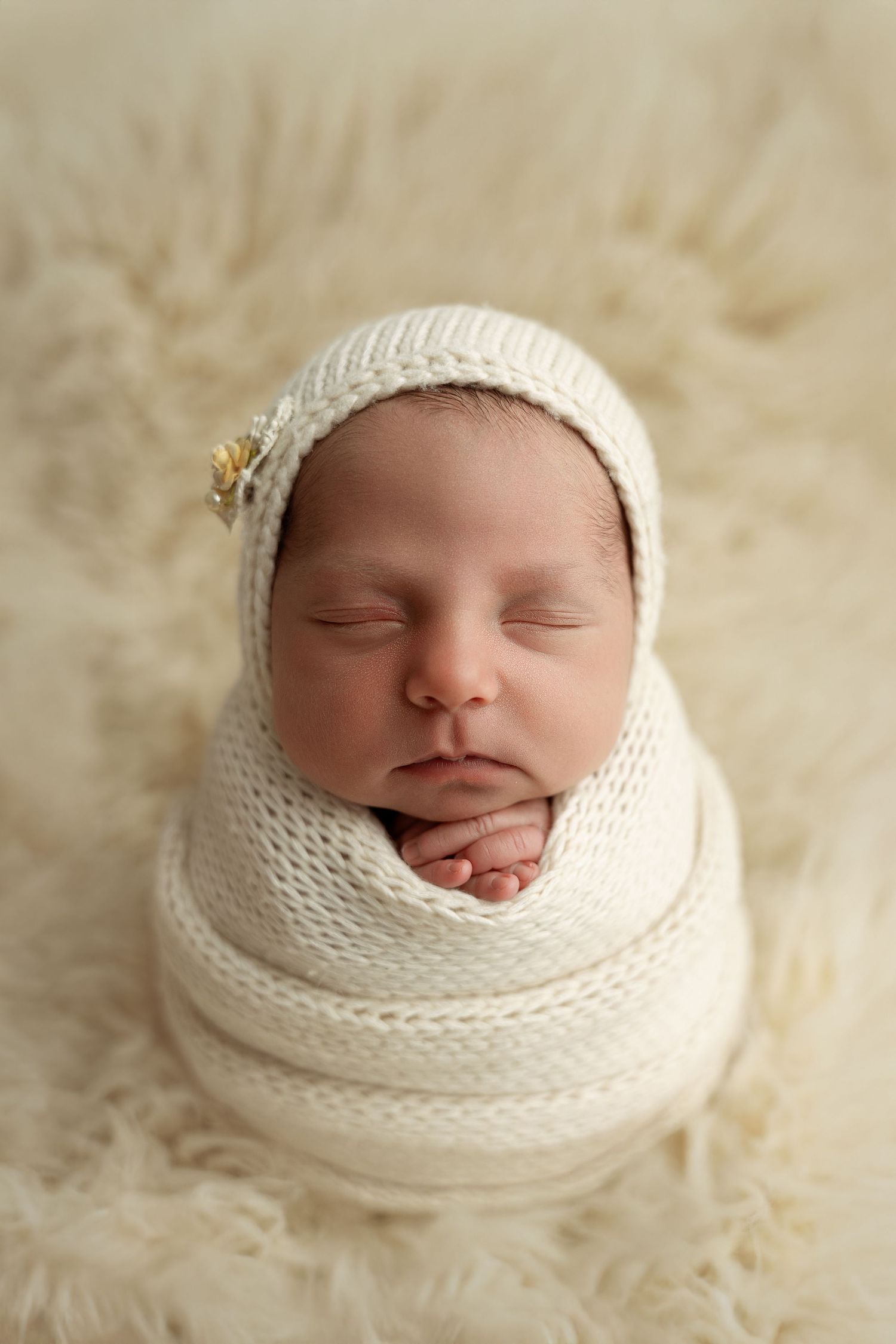 Sleeping newborn baby wrapped in cream knit swaddle with matching bonnet on fluffy blanket.