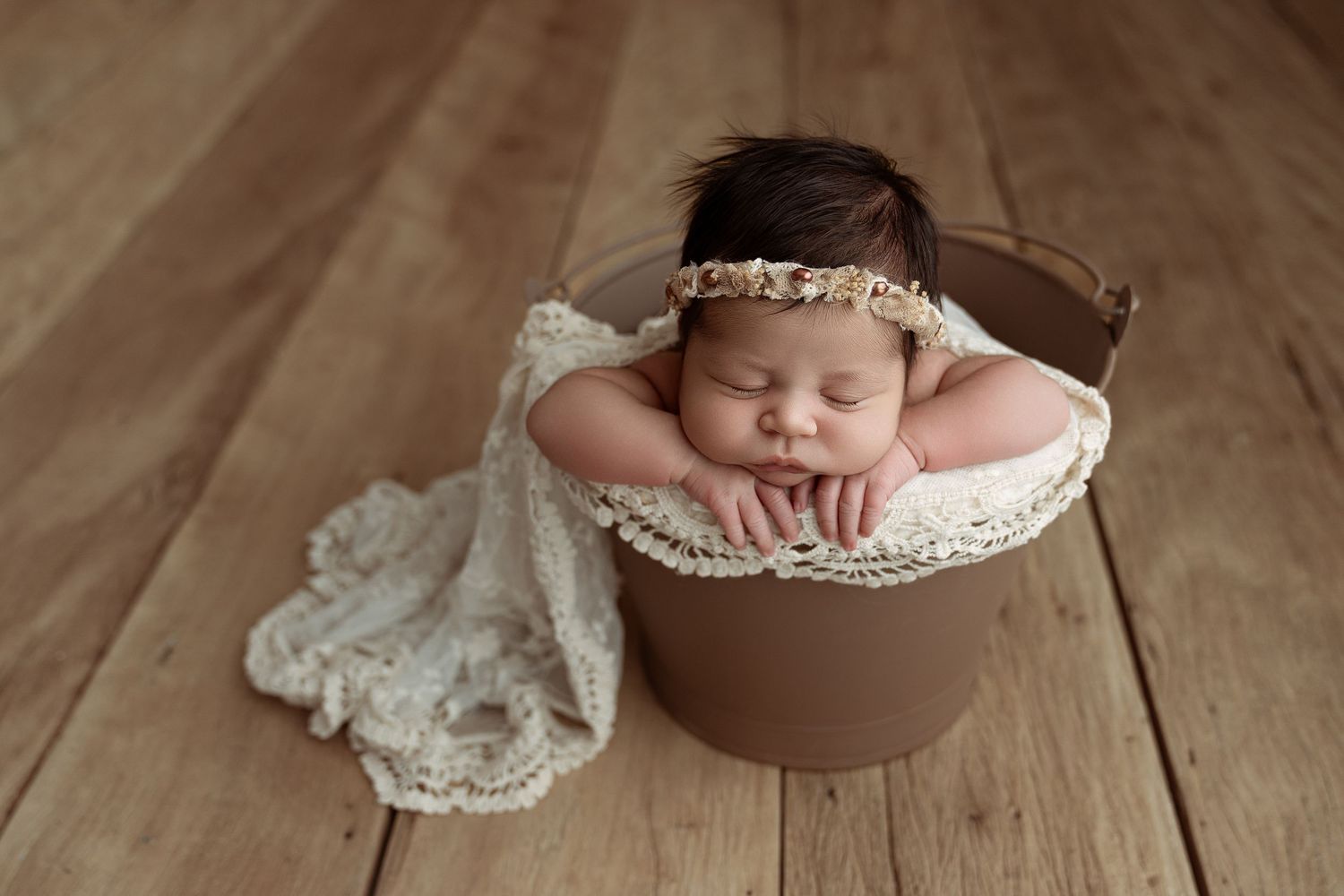 Newborn baby sleeping peacefully in a brown bucket with lace wrap and floral headband on wooden floor.