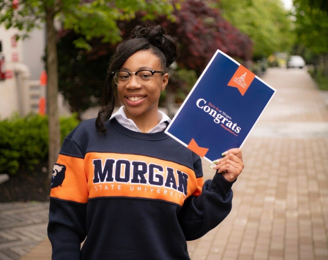 Happy graduate in Morgan State University sweatshirt holding a Congrats sign on a tree-lined path.