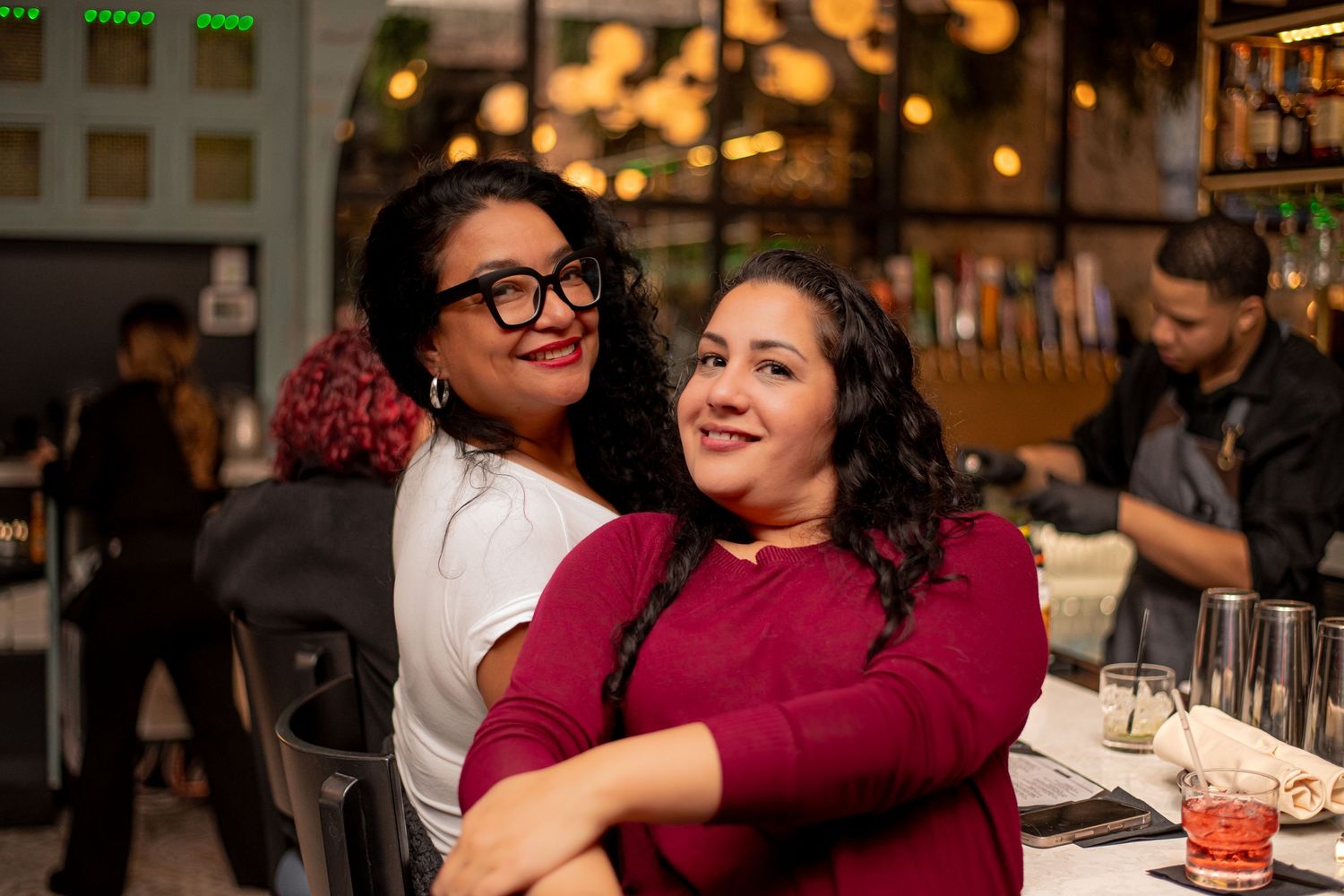 Two women smiling and embracing at a restaurant table with warm bokeh lighting behind them.
