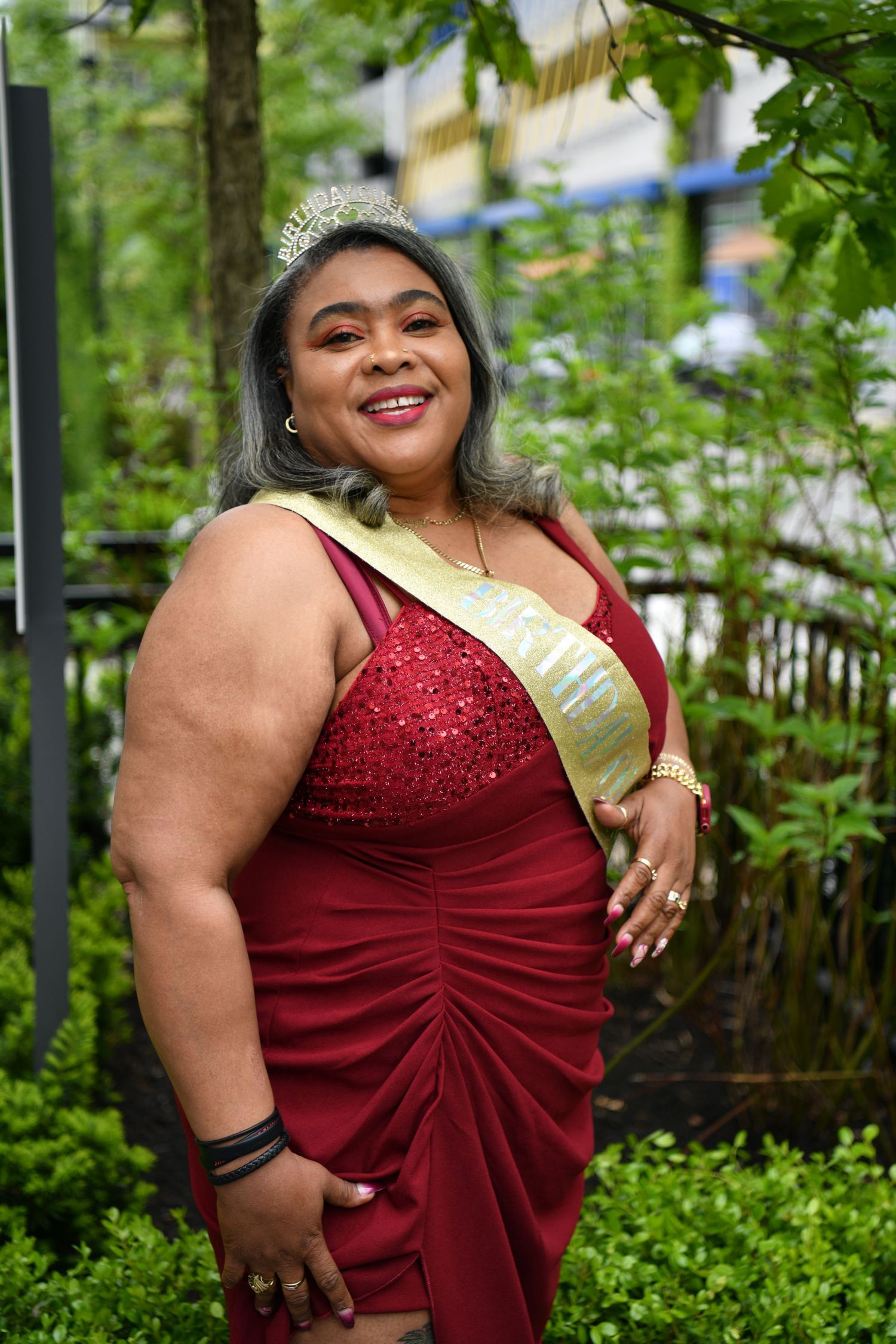 Smiling woman in a red sequin gown and tiara wearing a gold sash poses outdoors among green trees.
