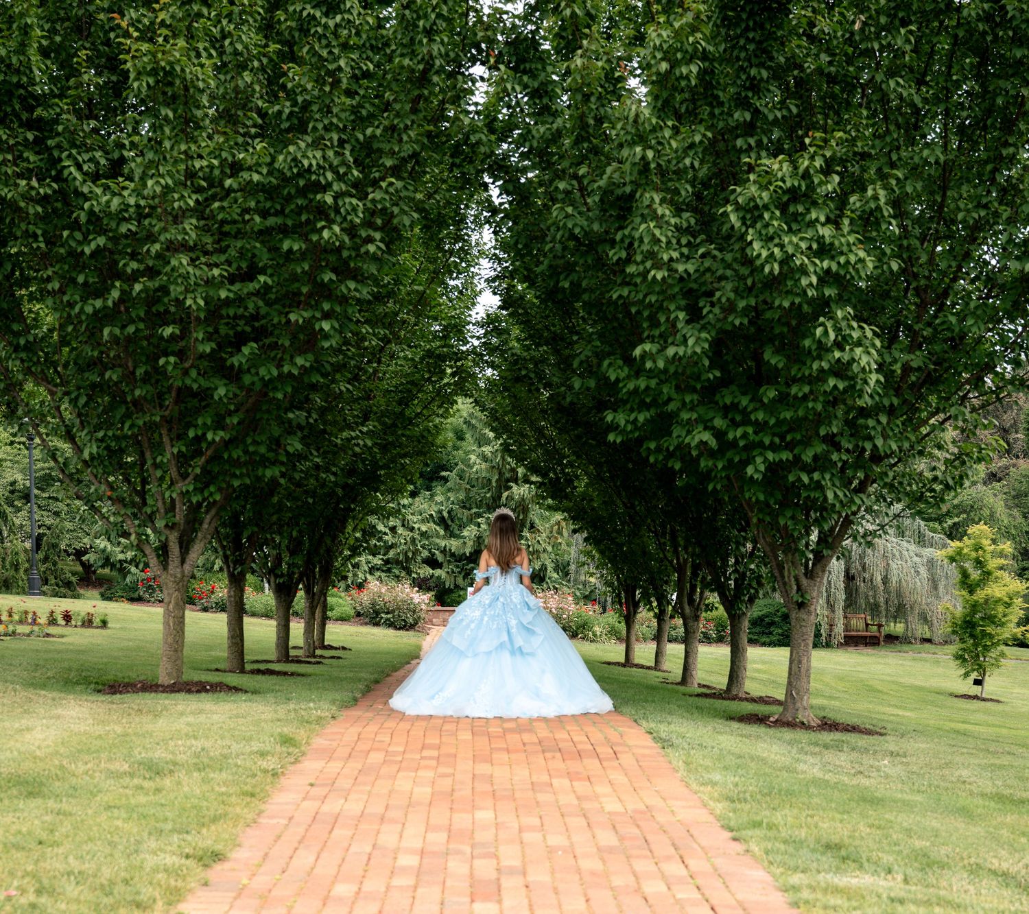 A girl in a light blue ball gown walks down a brick path lined with tall green trees in a lush garden.