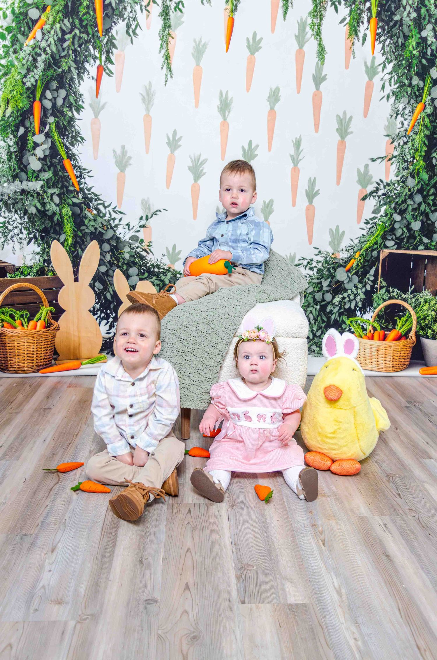 Three young children pose in an Easter-themed photo with carrot backdrop, bunny decor, wicker baskets, and stuffed animals.