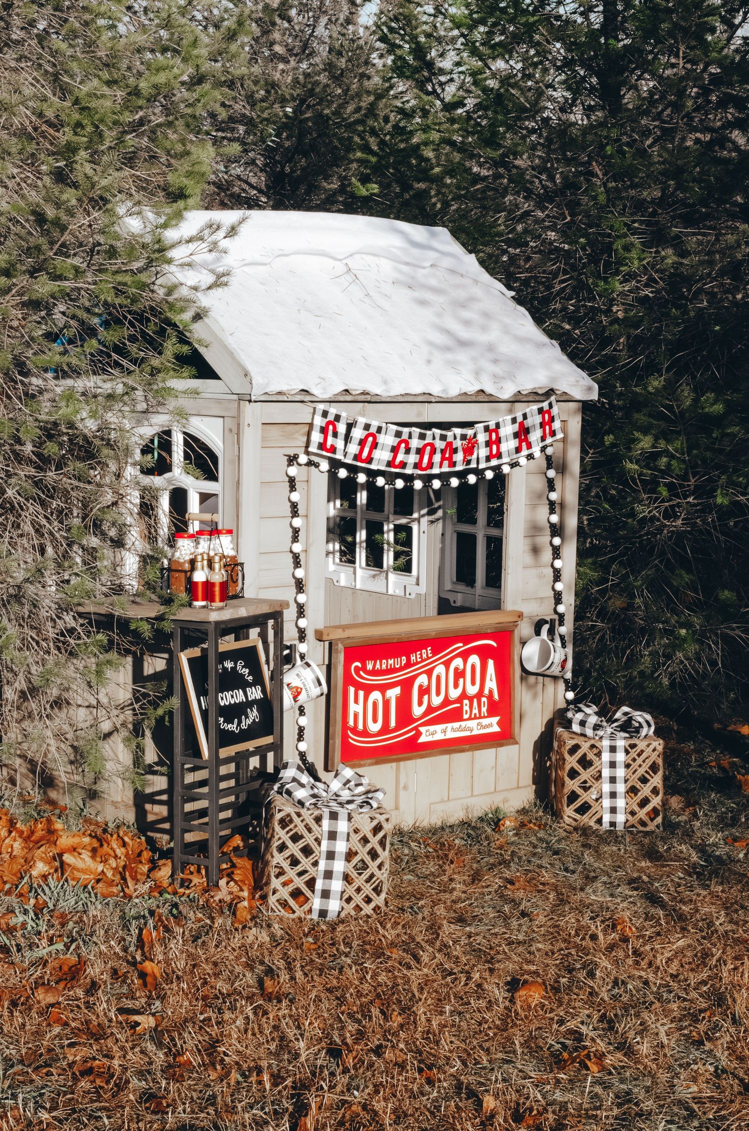 A charming outdoor hot cocoa bar stand decorated with Christmas lights, buffalo plaid gift boxes, and a red sign in a winter setting.