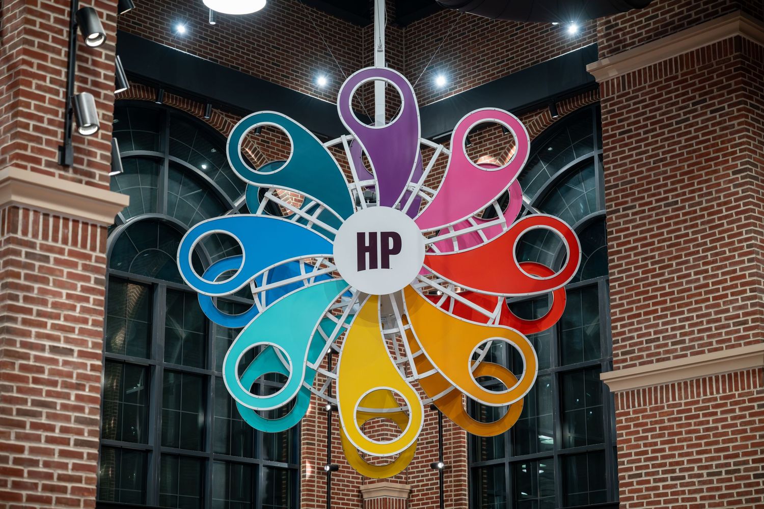 Colorful HP logo sign with rainbow petals and location pins displayed indoors against a brick wall backdrop.