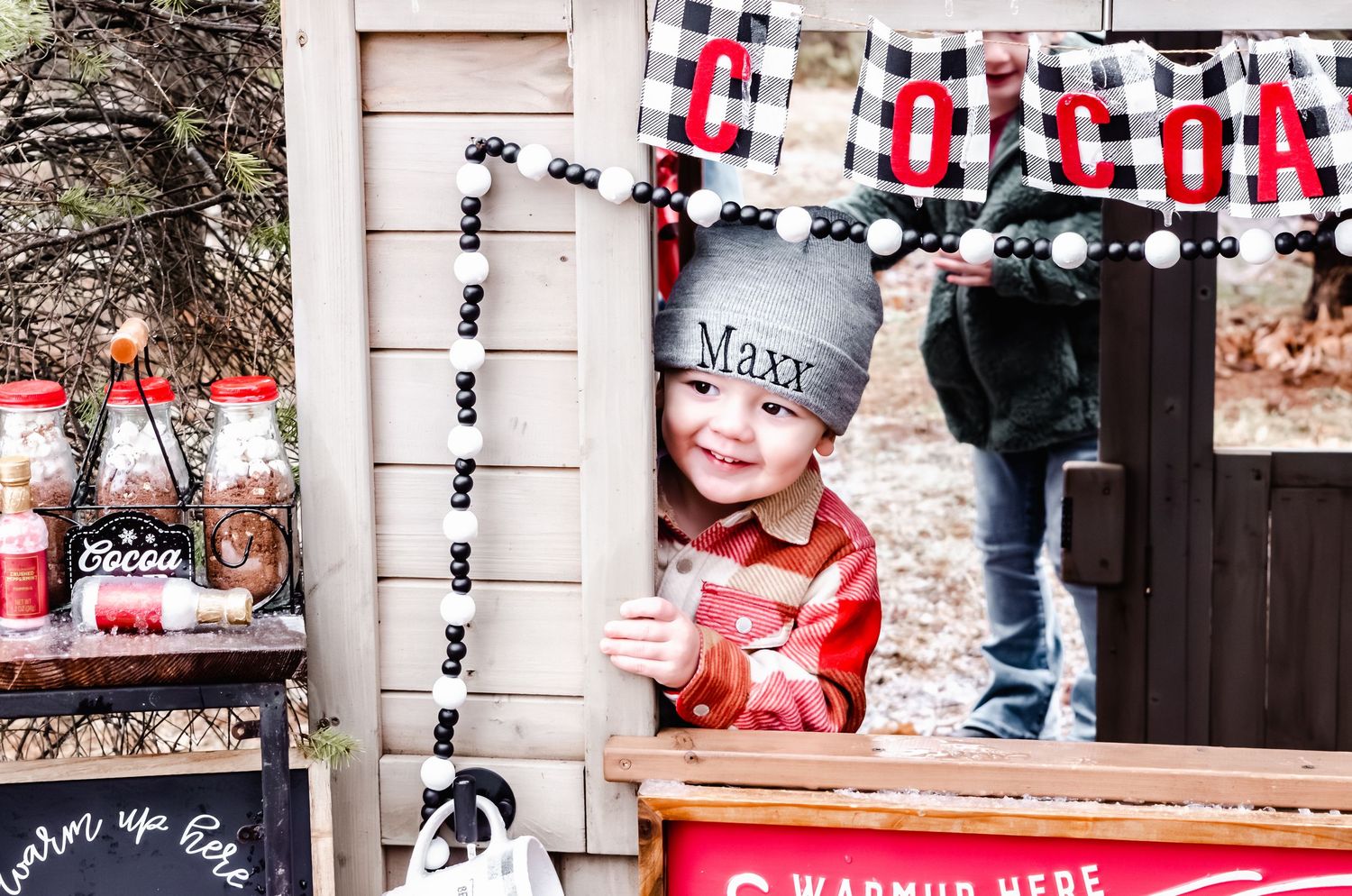 A smiling toddler named Maxx peers from a rustic cocoa stand, wearing a gray beanie and red flannel shirt.