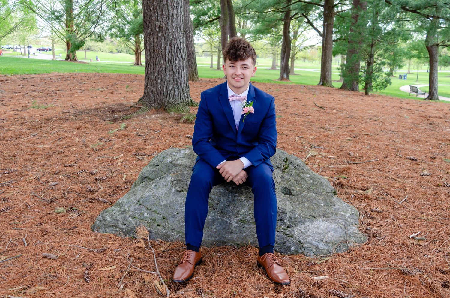 Young person in blue suit sitting on rock outdoors among pine trees, wearing brown oxford shoes and floral boutonniere.