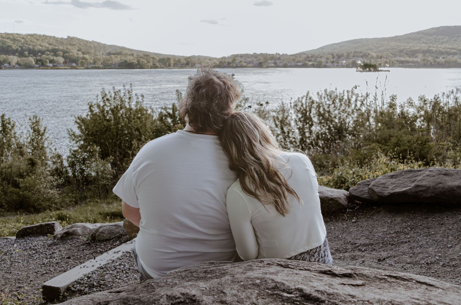 Couple sitting together on rocks overlooking a serene lake surrounded by lush green trees and hills.