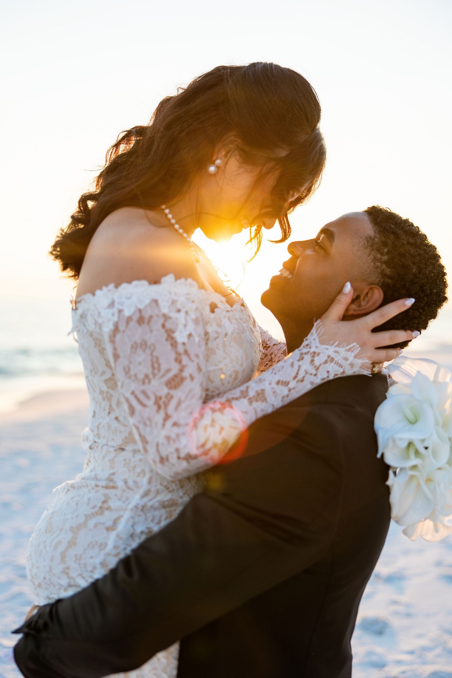 A bride and groom share a romantic embrace on a beach at sunset, with golden light glowing behind them.