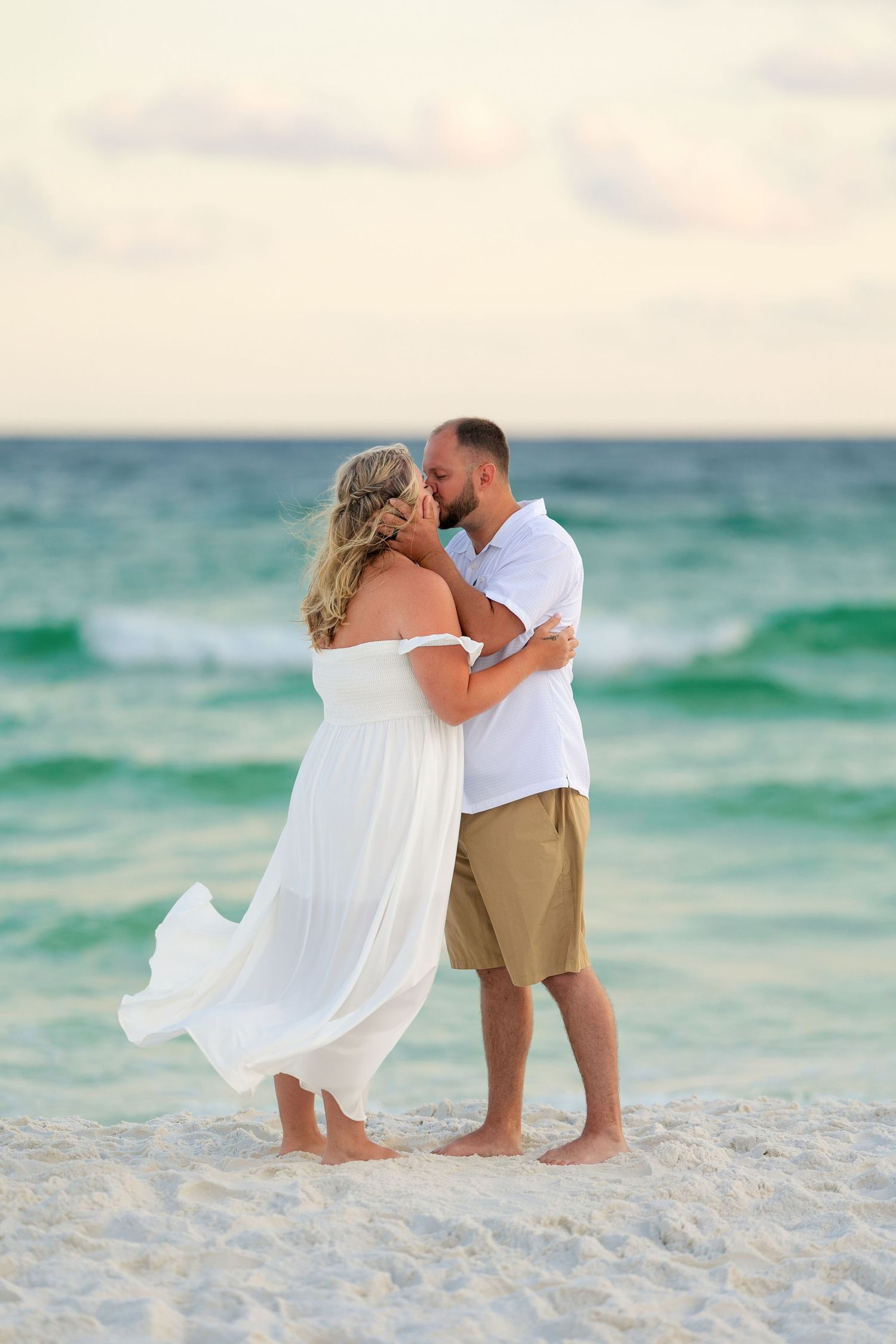 Couple kissing on a white sand beach, woman in flowing white dress, man in khaki shorts, turquoise ocean waves behind them.