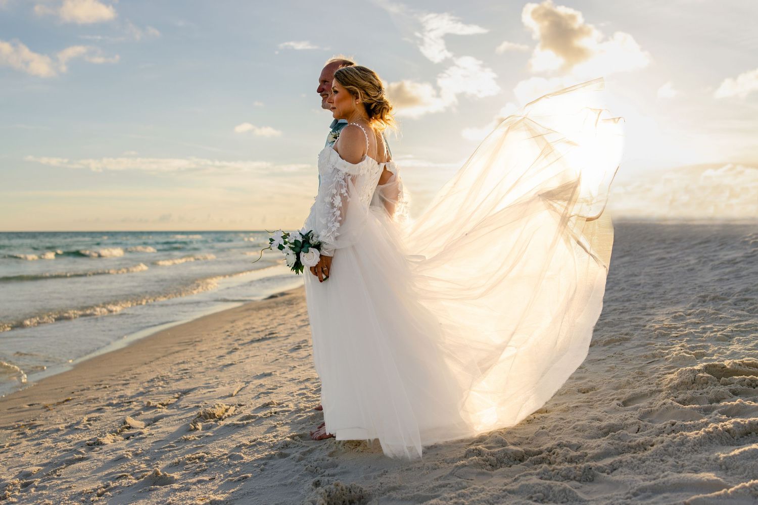 Bride in flowing white lace gown with veil blowing in wind on sunny beach holding bouquet at sunset.