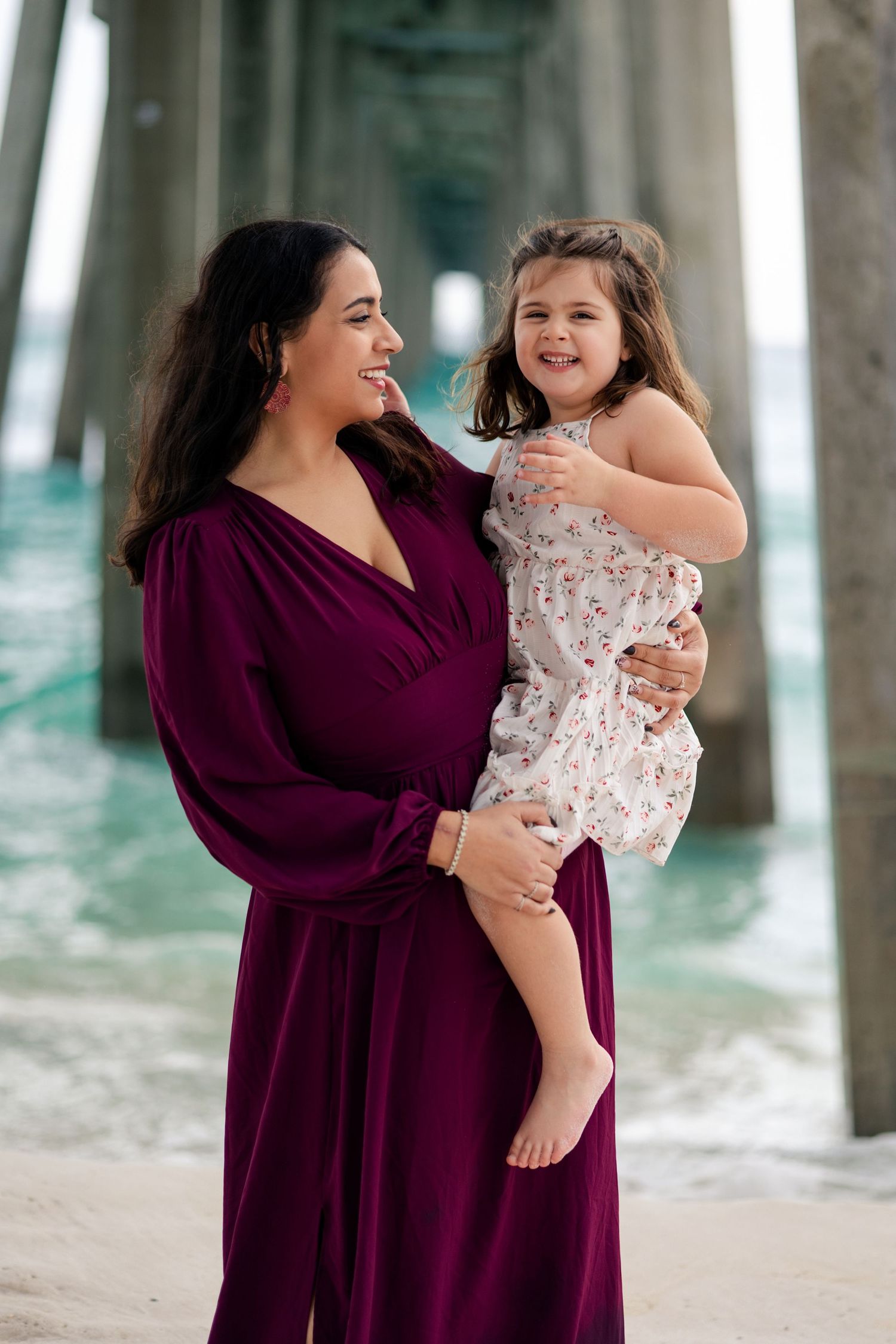 A woman in a burgundy dress holds a smiling toddler girl in a floral dress under a beach pier.