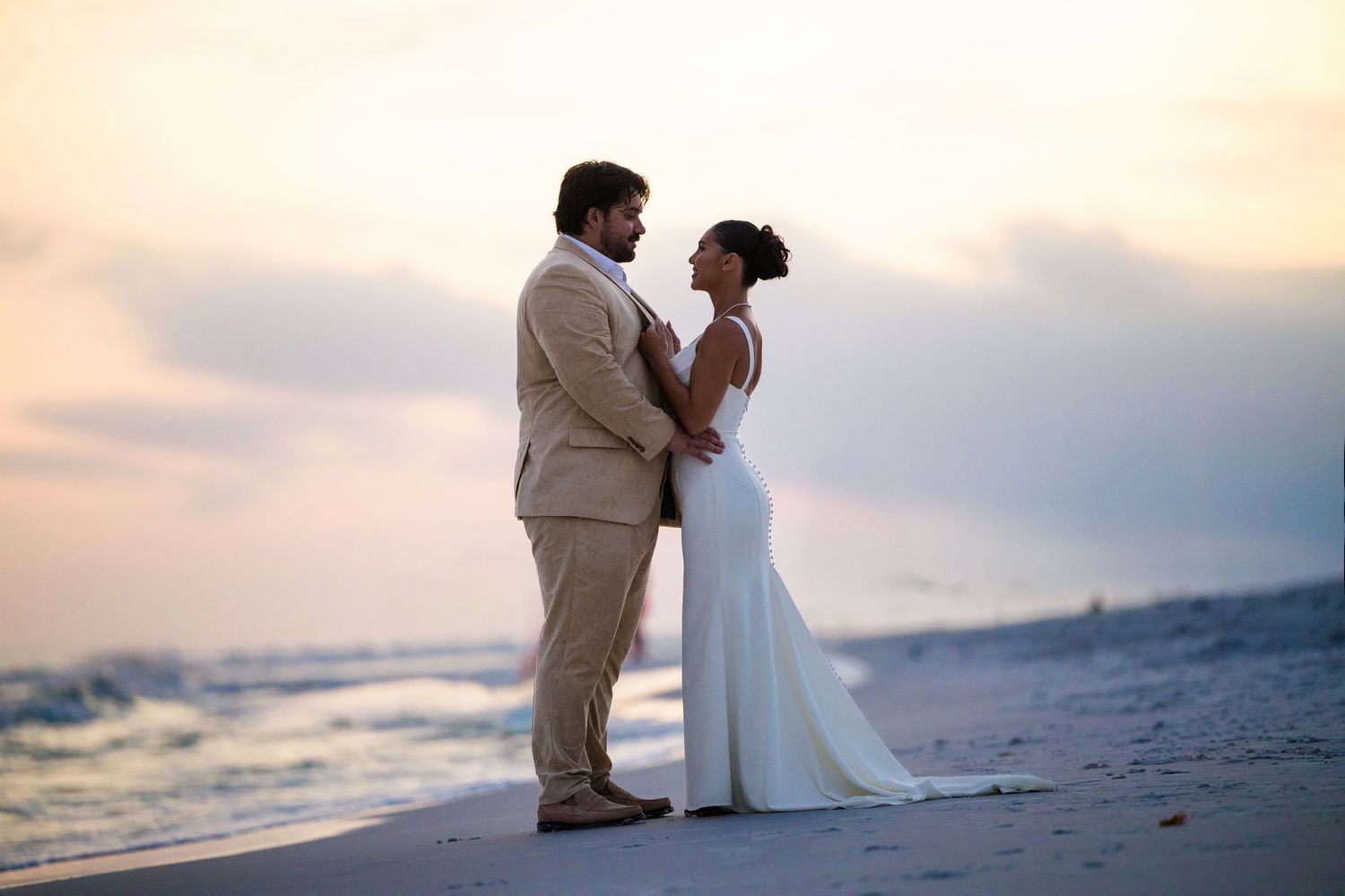 Bride and groom share a romantic moment on a beach at sunset, with ocean waves in the background.