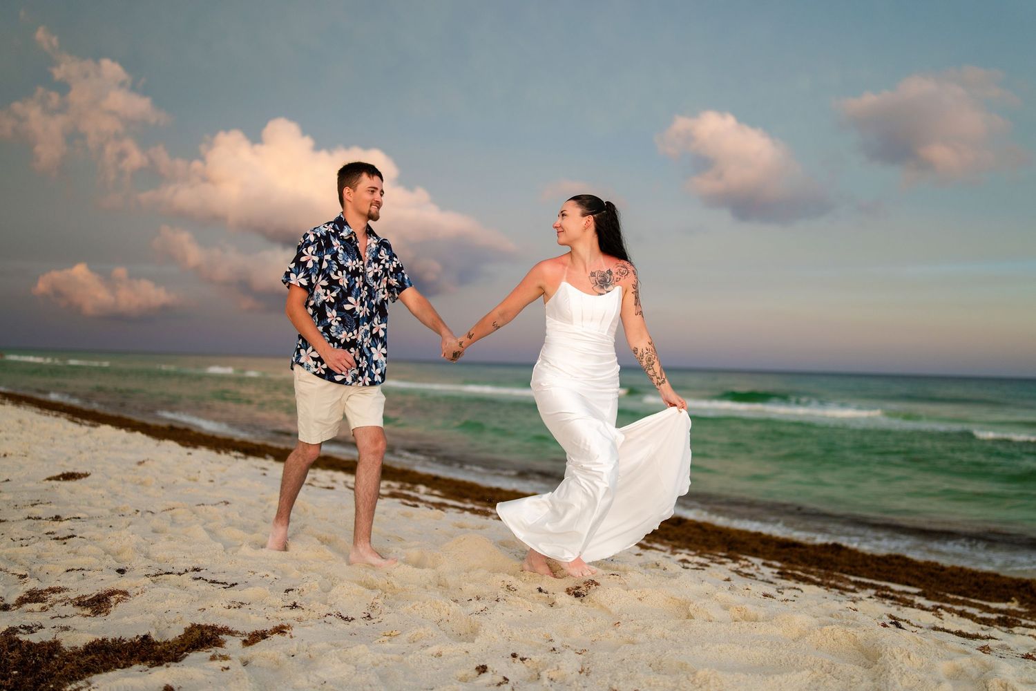 Couple in wedding attire holding hands and running joyfully on a sandy beach at sunset.