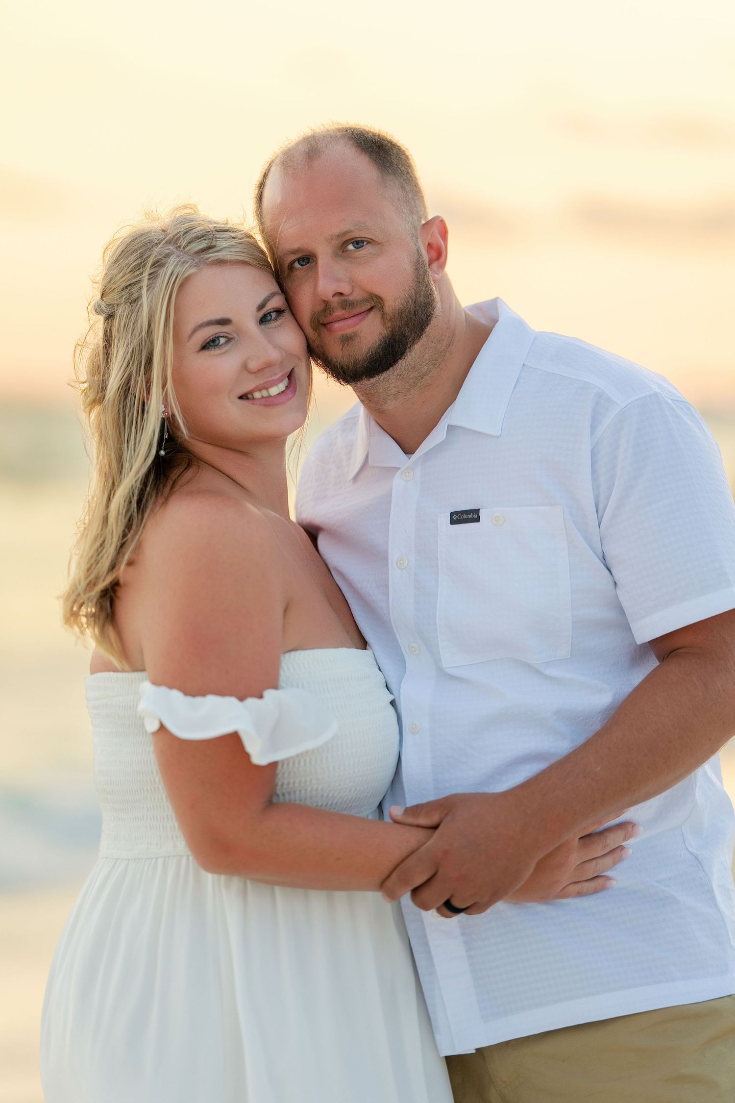 A smiling couple in white outfits embracing on a beach at sunset, woman in off-shoulder dress, man in button-up shirt.
