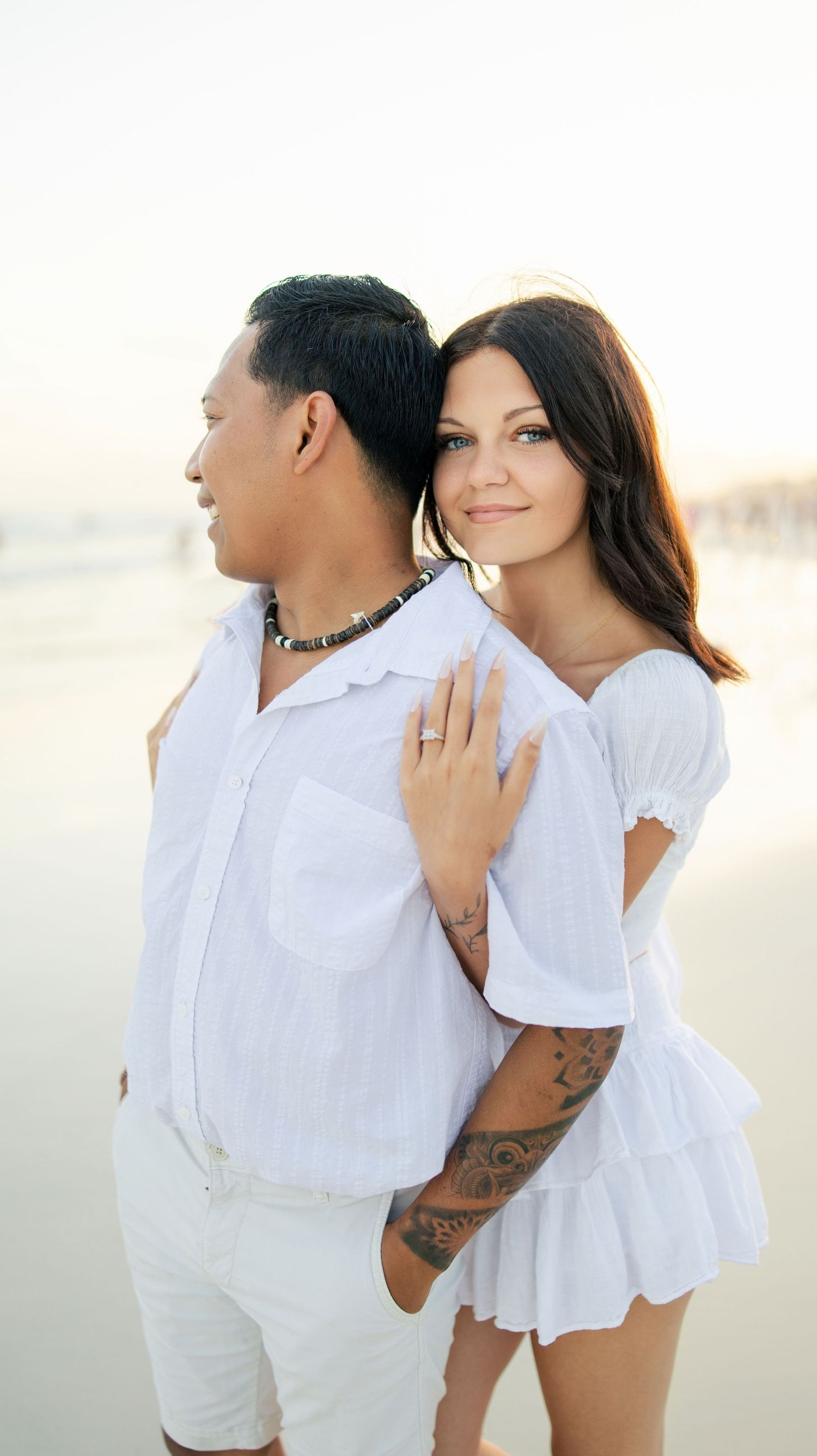 Couple in matching white outfits embracing on a beach at sunset, woman smiling with tattoos visible.