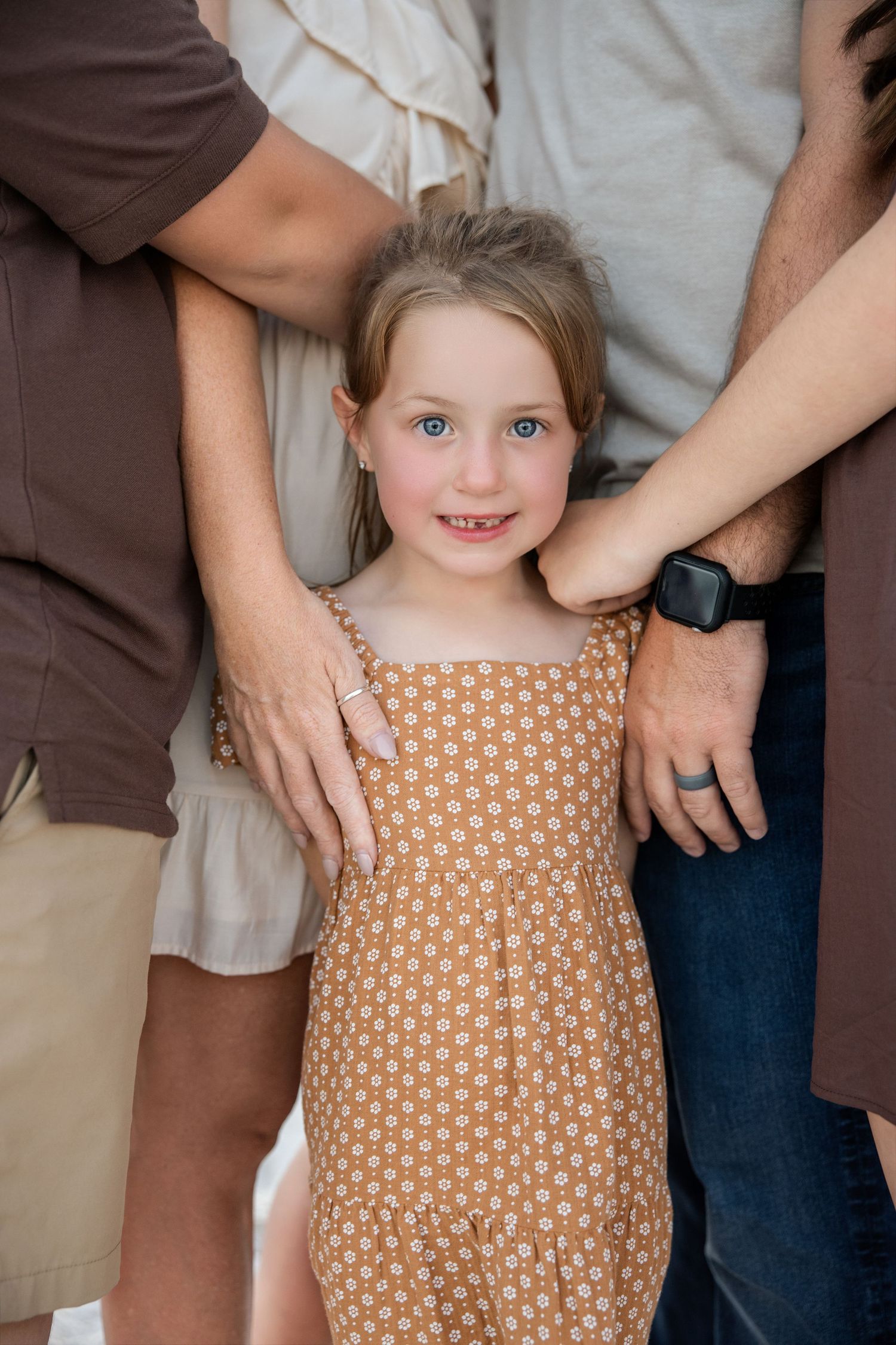 Young girl in floral dress smiling at camera, flanked by parents hands on her shoulders.