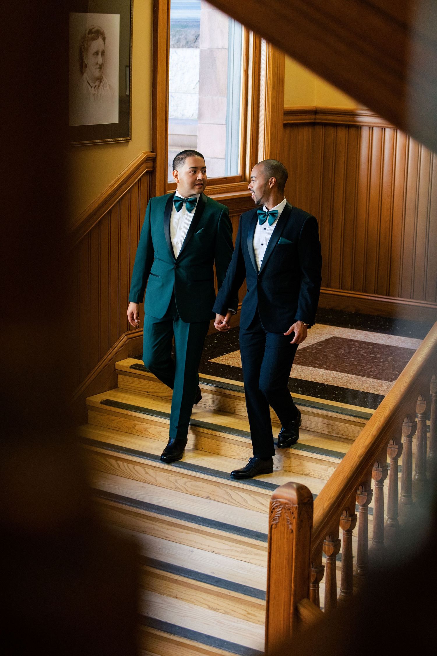 Two grooms in teal and navy tuxedos hold hands walking down elegant wooden staircase at wedding venue.