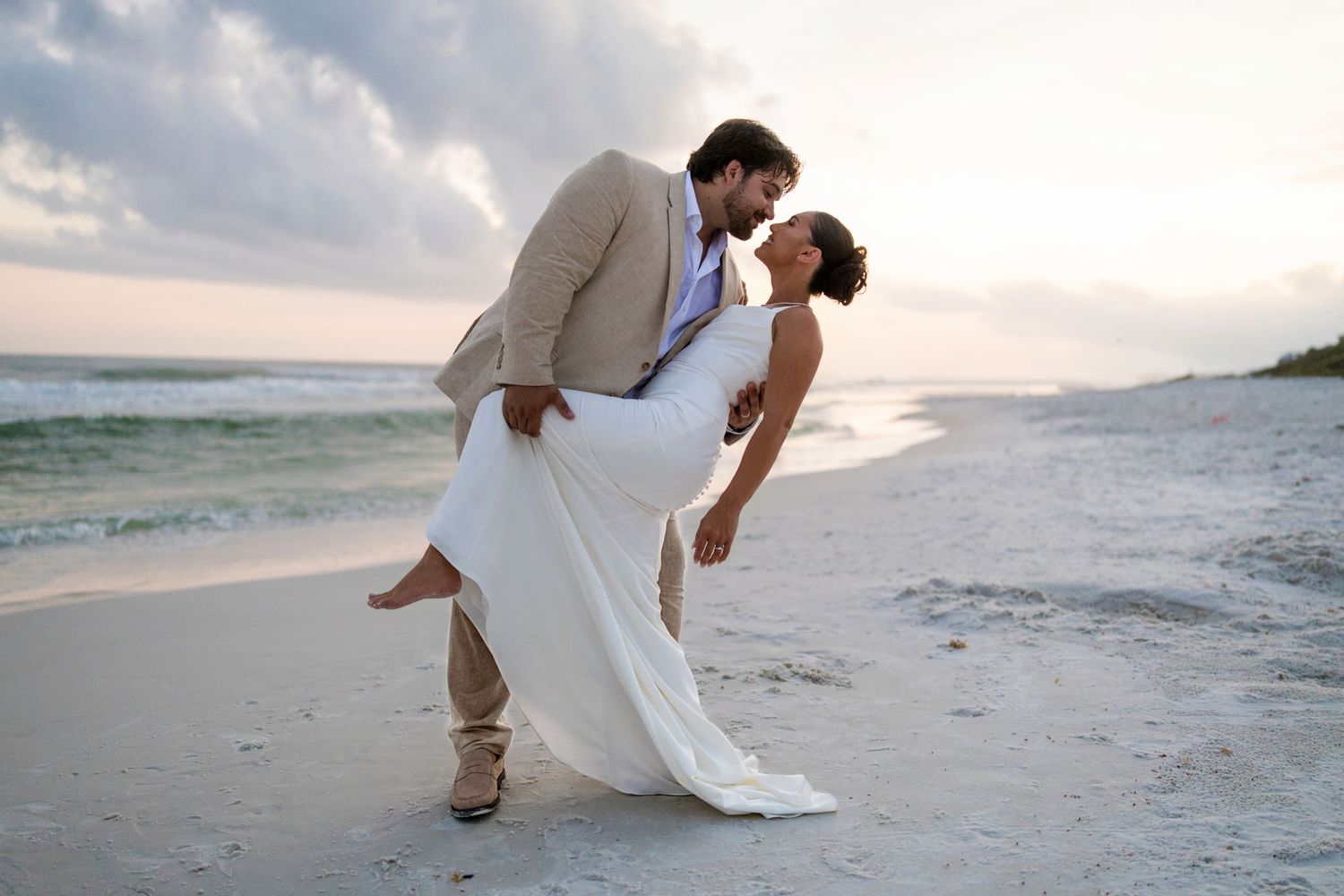 Couple sharing a romantic dip kiss on a sandy beach at sunset, groom in tan suit, bride in white wedding gown.