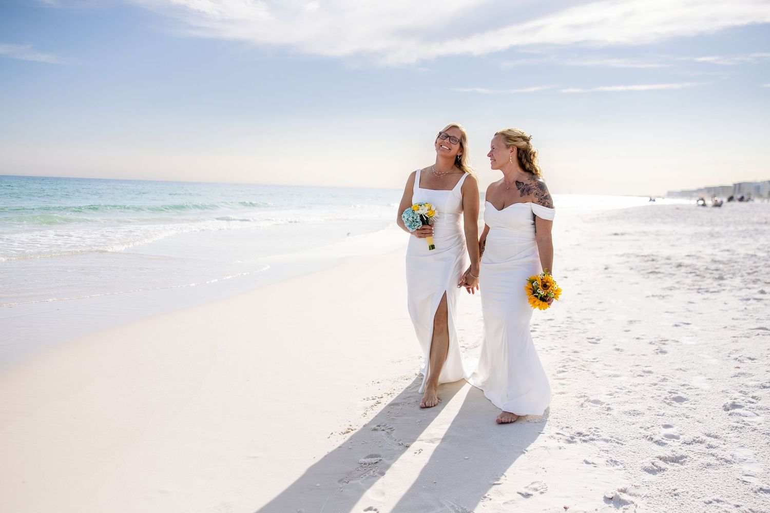 Two brides in white dresses holding sunflower bouquets walk hand in hand on a sunny white sand beach.