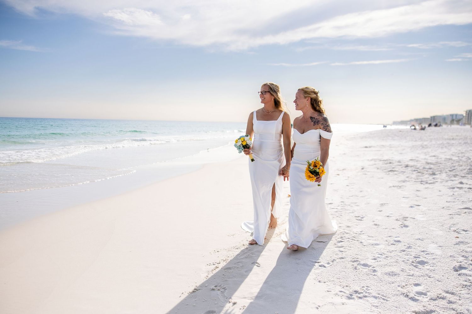 Two brides in white gowns holding sunflower bouquets walk barefoot on a sunny beach near the ocean.