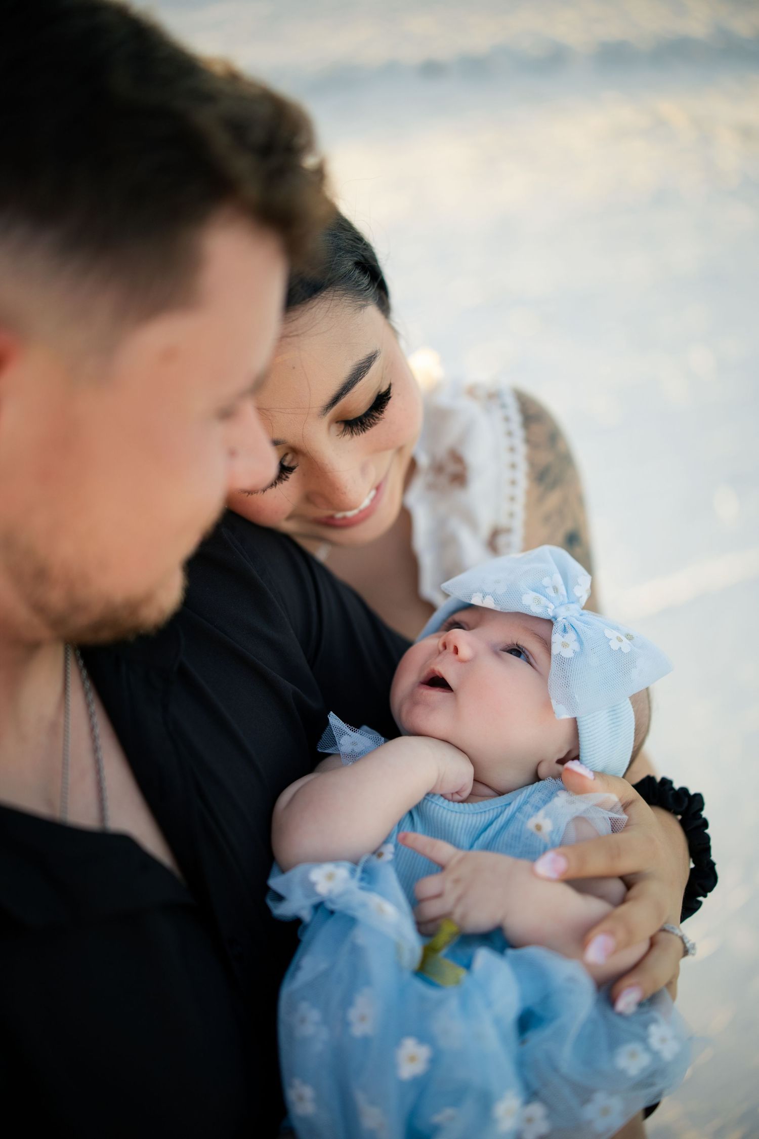 Young couple smiling and holding their newborn baby dressed in blue with a bow headband outdoors.
