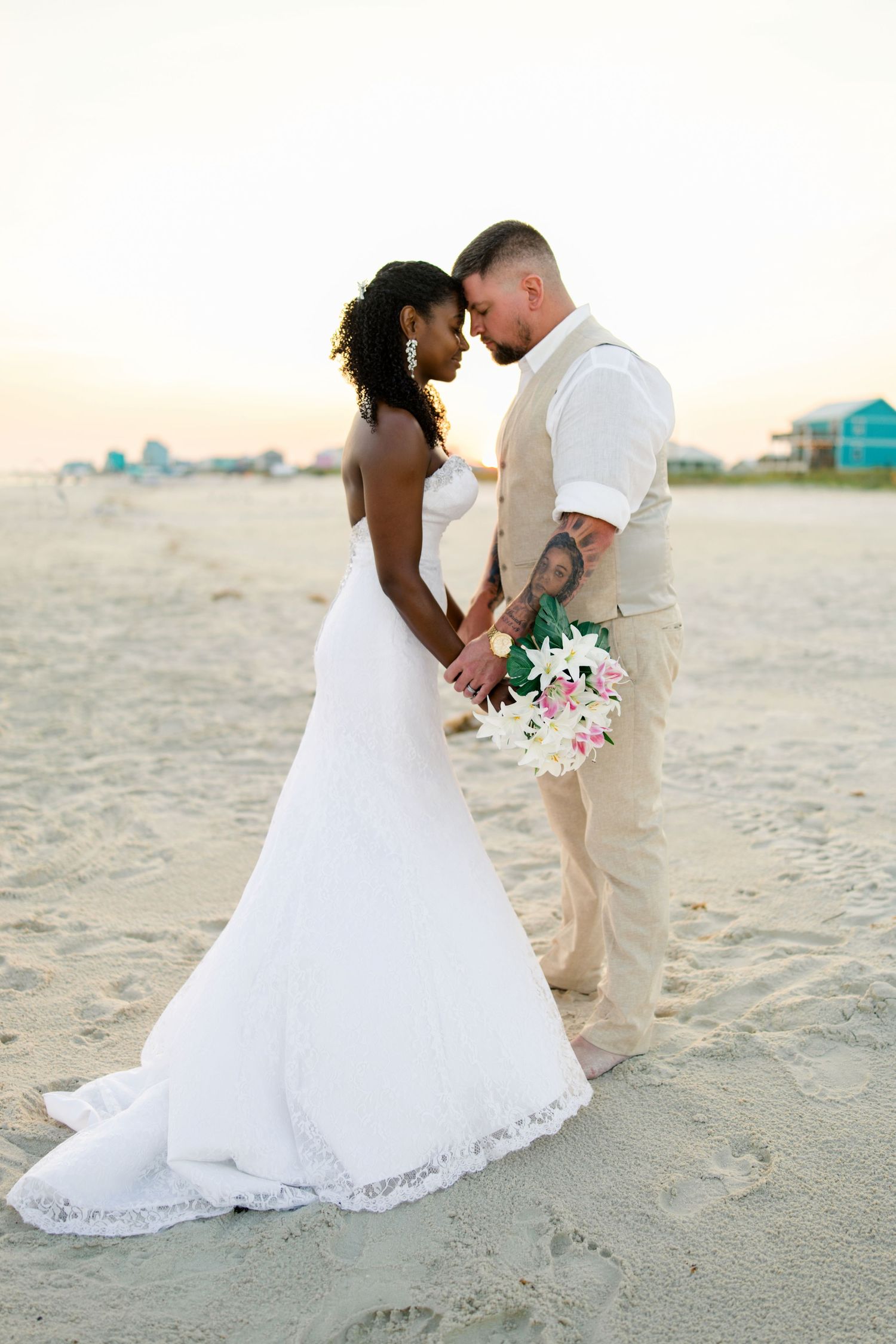 Interracial couple in wedding attire holding hands on a sandy beach at sunset, bride in white lace gown holding pink bouquet.