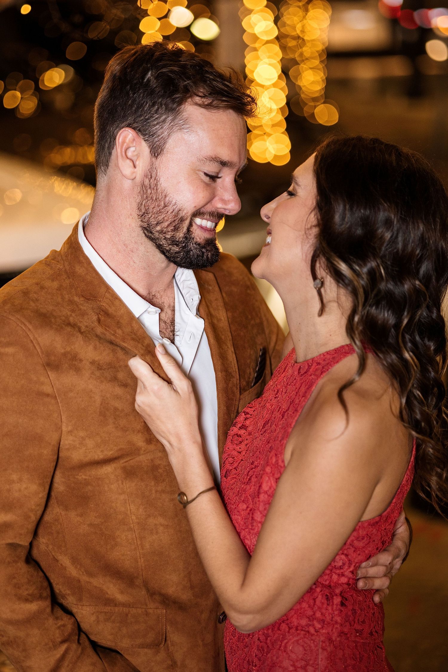 A smiling couple in elegant attire share a romantic moment with bokeh lights in the background.