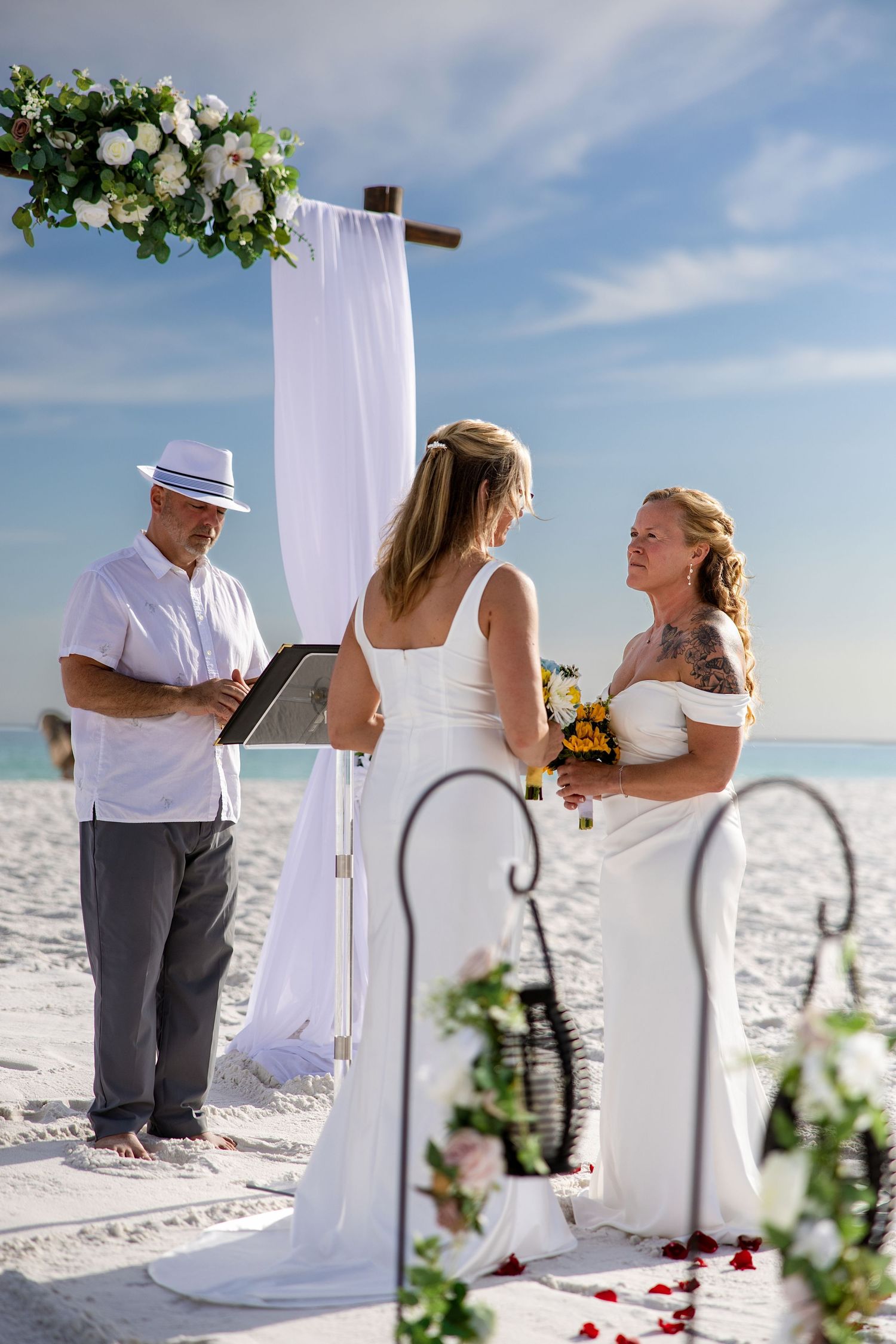 Two brides exchange vows at a beach wedding ceremony under a floral arch, officiated by a man in a white hat.