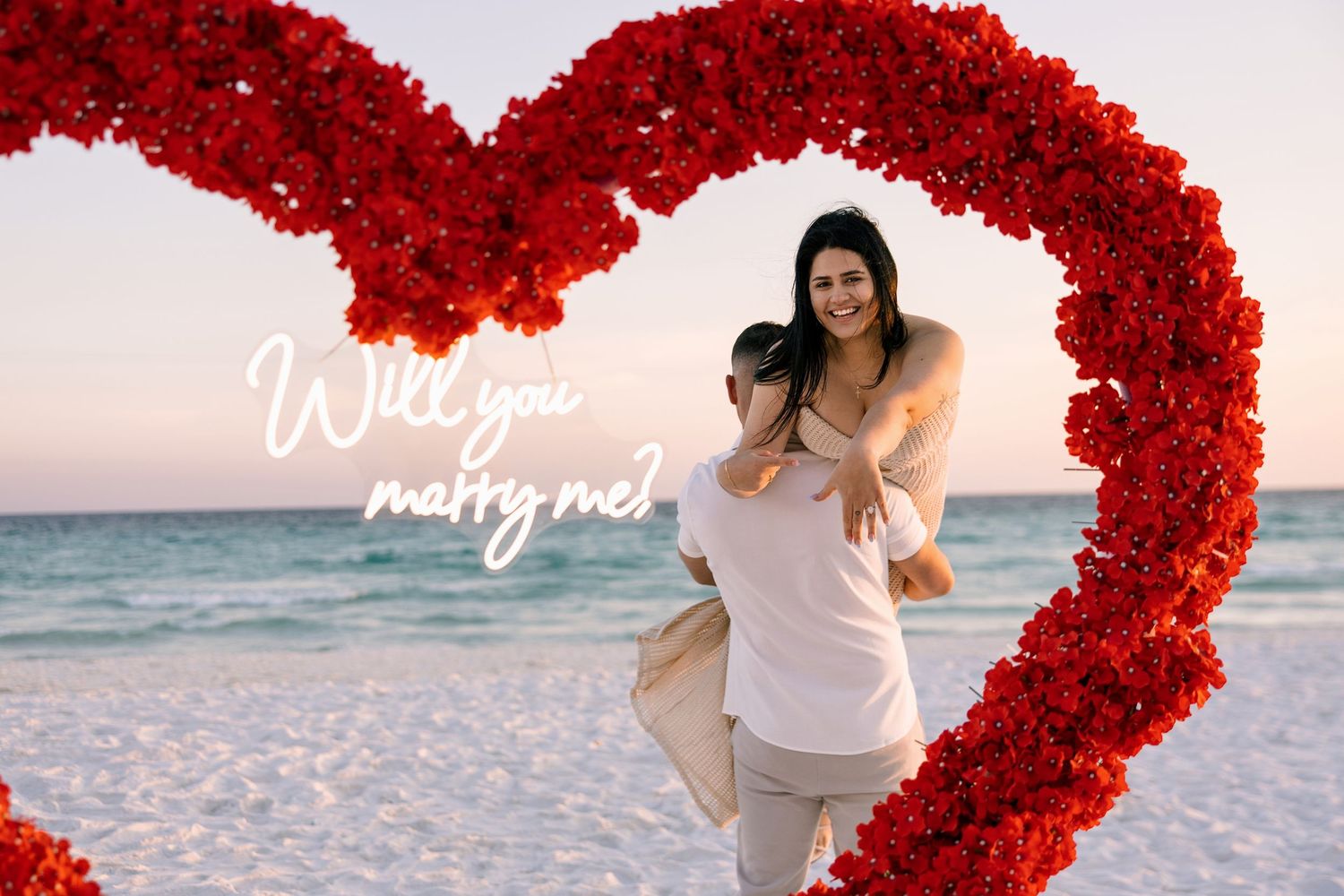 Couple embracing on a beach framed by a red rose heart arch with Will You Marry Me text at sunset in pensacola, florida