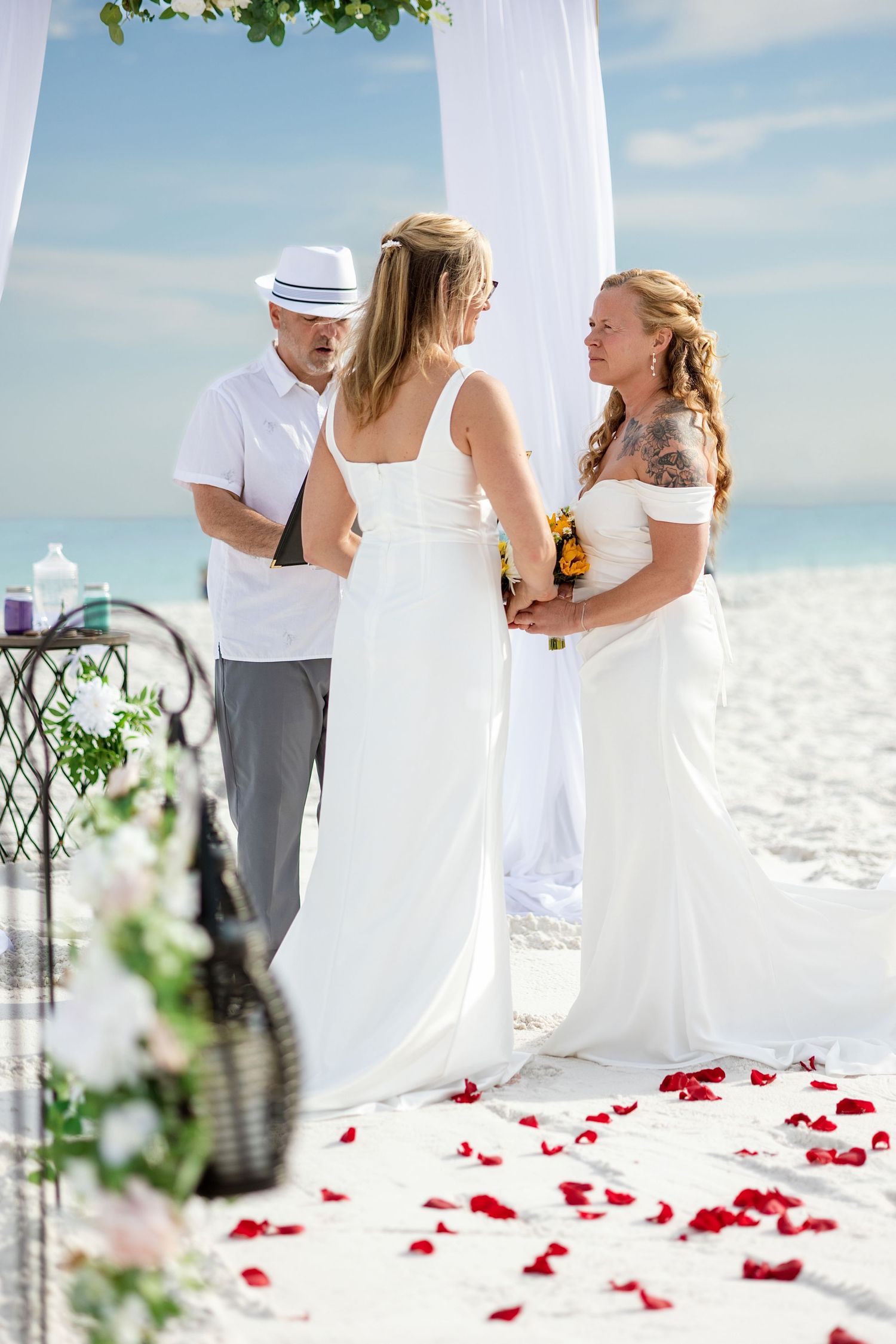 Two brides in white dresses exchange vows at a romantic beach wedding ceremony with rose petals on the aisle.